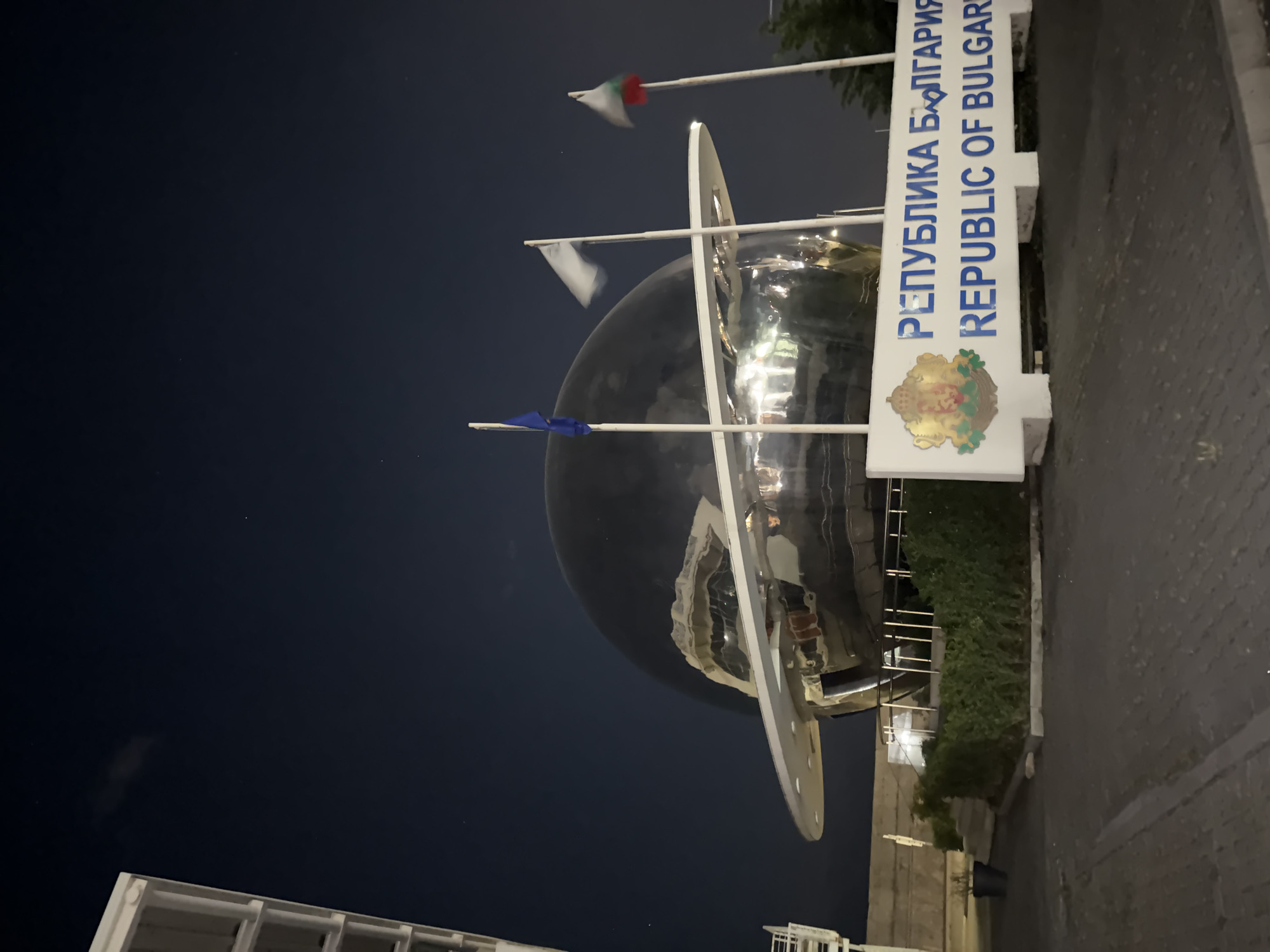 This image shows a monument in Burgas, Bulgaria. It features a chrome sphere with three rings around it and flags extending from the sphere. A sign reads "Republica Bulgaria - Republic of Bulgaria" in both Bulgarian and English. The monument is located on a paved area, with some greenery nearby, and is photographed at night. The sky is dark, and the scene is lit artificially. There are buildings visible in the background.