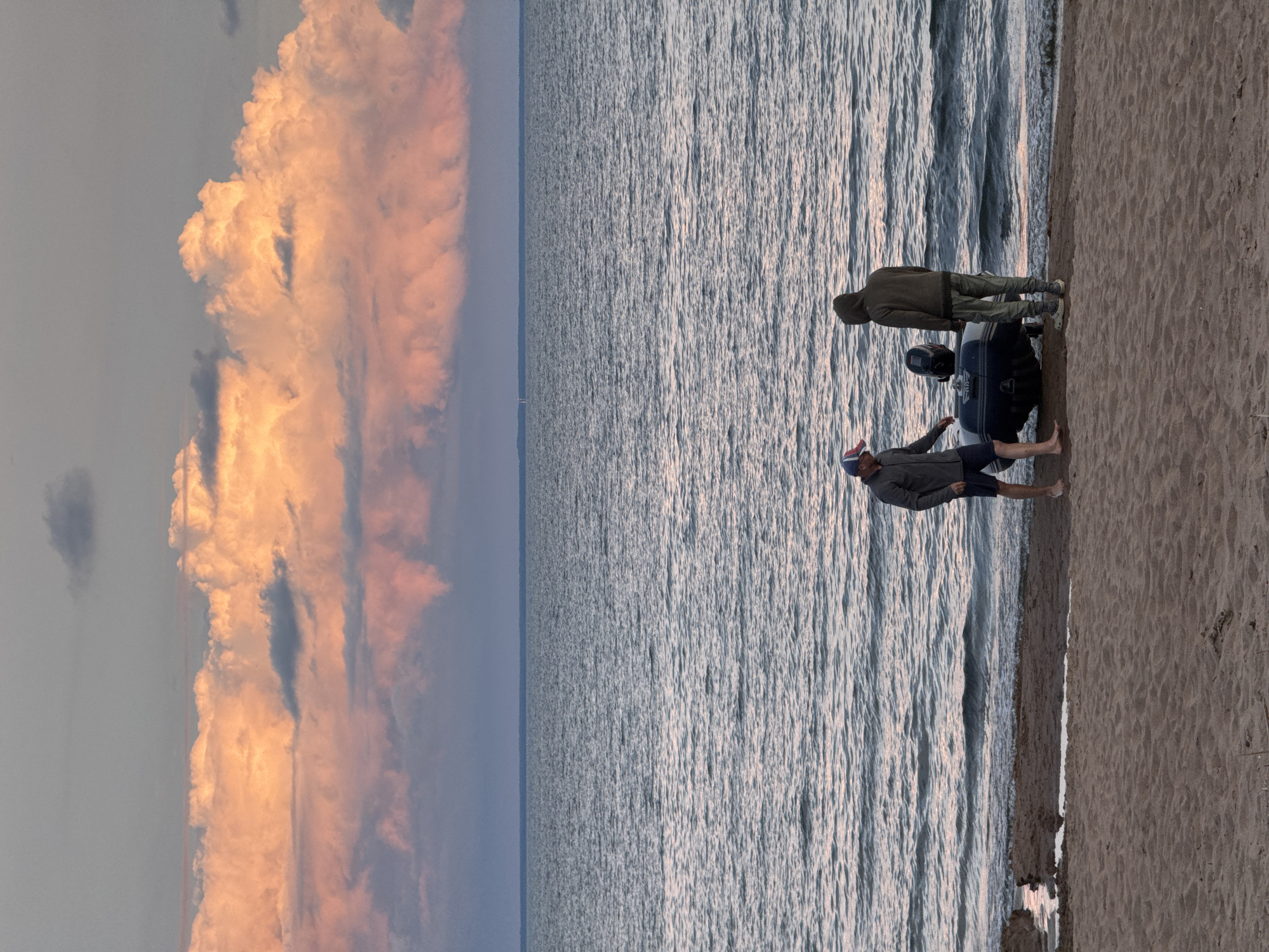 Two individuals are present on a sandy beach adjacent to a calm body of water. One person, wearing a blue baseball cap, dark shorts, and a grey jacket, stands near the water's edge, facing a deflated inflatable boat with an outboard motor. The second individual, wearing a hooded green jacket and patterned pants, stands behind the boat, also facing it. The sky above is dominated by large, dramatic clouds illuminated with shades of pink and orange, suggesting either sunrise or sunset. The water is a deep blue with gentle ripples, and the horizon line is visible in the distance. The location is identified as Shepelevo, Russia.