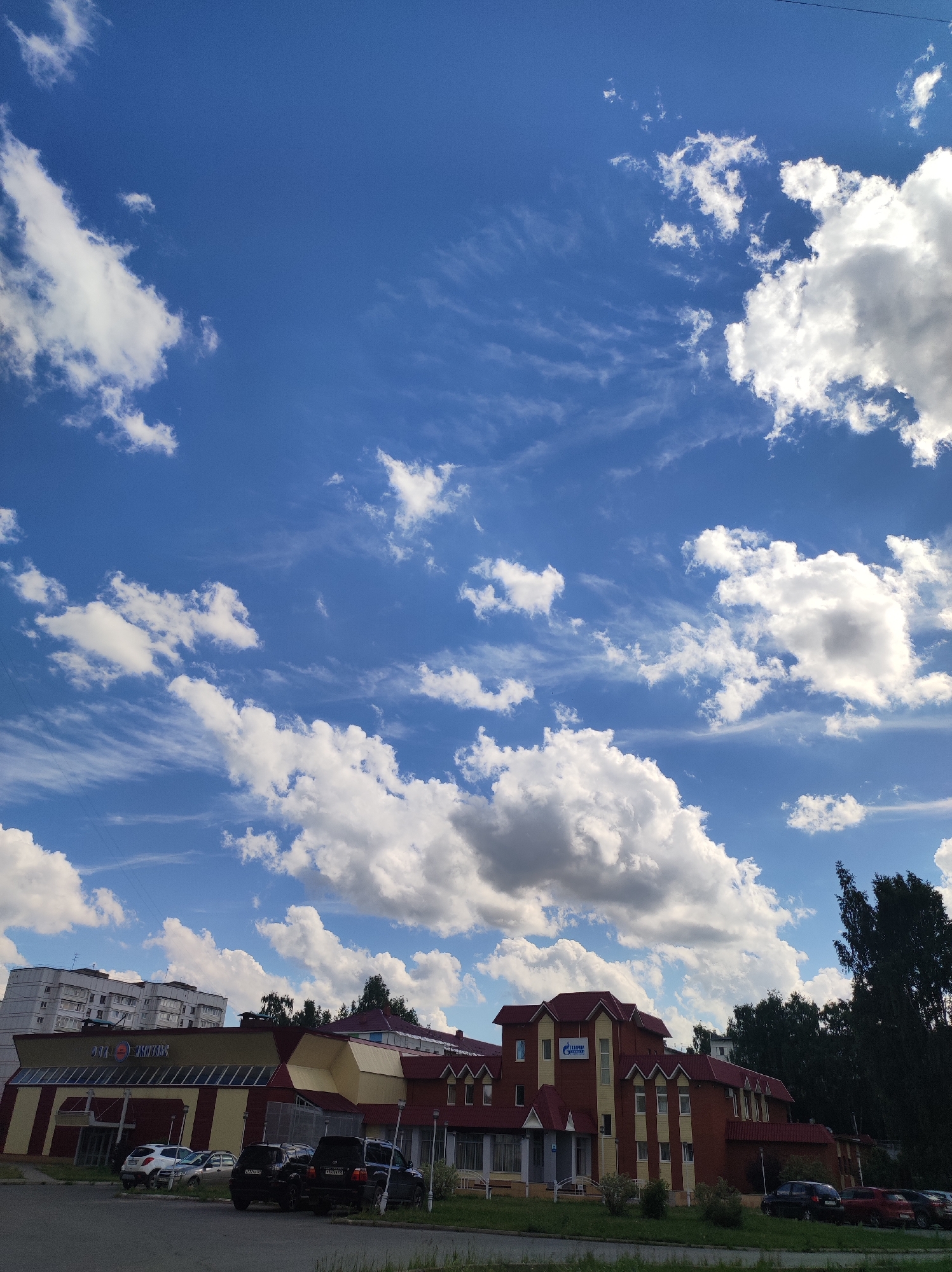 The image captures a scene in the city of Chaikovsky, Russia, featuring a bright blue sky with scattered white cumulus clouds. In the foreground, a paved area with parked cars leads to a row of buildings. The buildings appear to be a commercial complex, with a prominent yellow and red structure on the left, and a taller, red-roofed building with yellow accents on the right. The building on the left has large windows and signage that is partially visible. The building on the right displays the Gazprom logo. Trees and greenery are visible between and behind the buildings, and a multi-story residential building is seen in the background on the left. The overall impression is a sunny day in an urban or suburban environment.