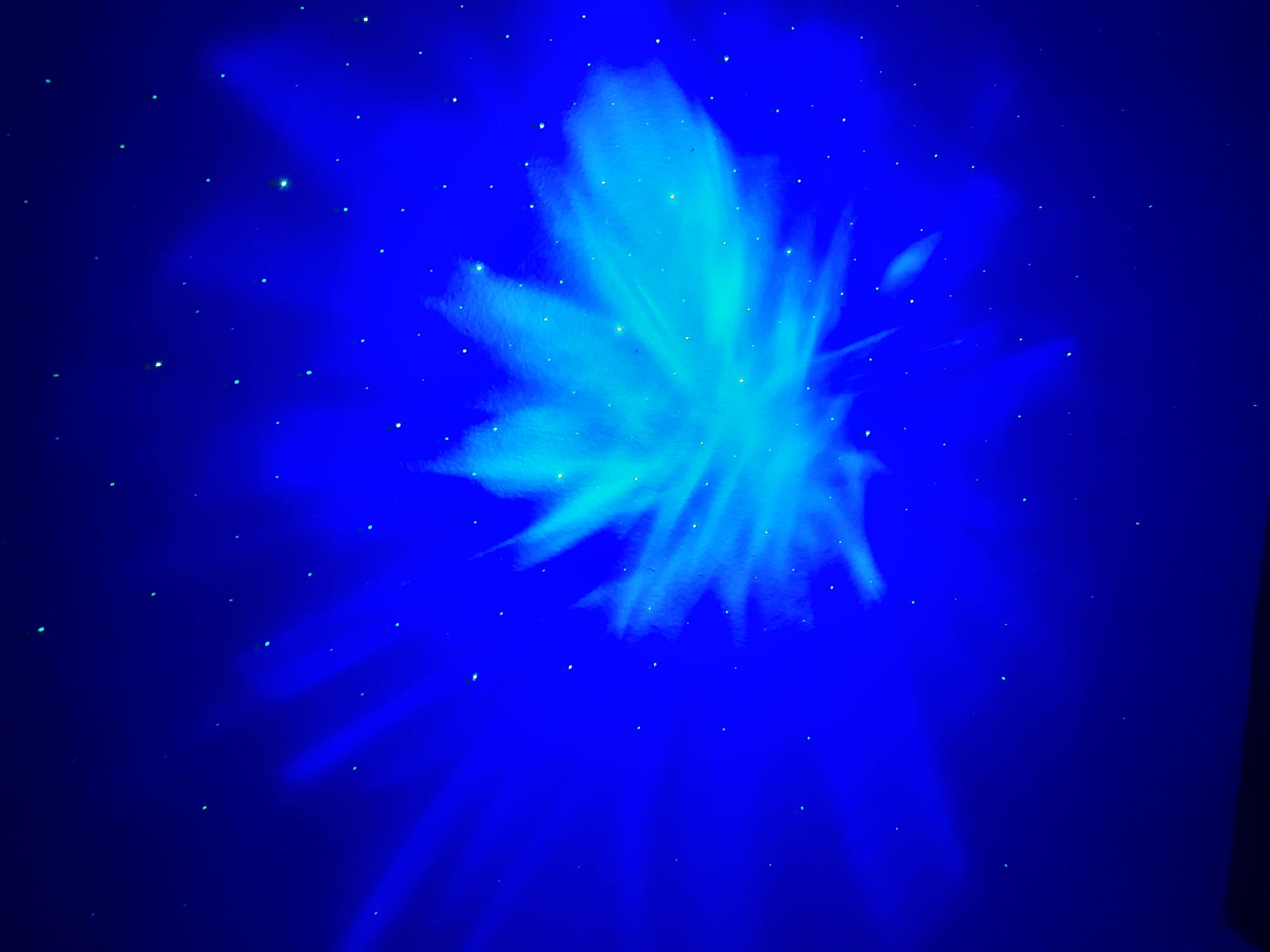 The image depicts a dark blue background illuminated by a bright, ethereal light projection in the center. The projection resembles a stylized flower or a burst of energy, with soft, flowing edges in shades of light blue and cyan. Scattered across the dark background are numerous small, bright specks, giving the impression of stars in a night sky or glowing particles. The overall mood is mystical and serene, with the strong contrast between the deep blue and the luminous light creating a dramatic effect. There are no visible people, objects, or text within the image. The setting appears to be an indoor or darkened outdoor space where light projections are being used. The time of day is likely night, given the dark backdrop and starry appearance. No weather or specific location cues are discernible, other than the provided location context of Baan Mae Nam, Thailand, which is not visually represented in the image itself. The activity taking place is the display of a light art installation or projection.