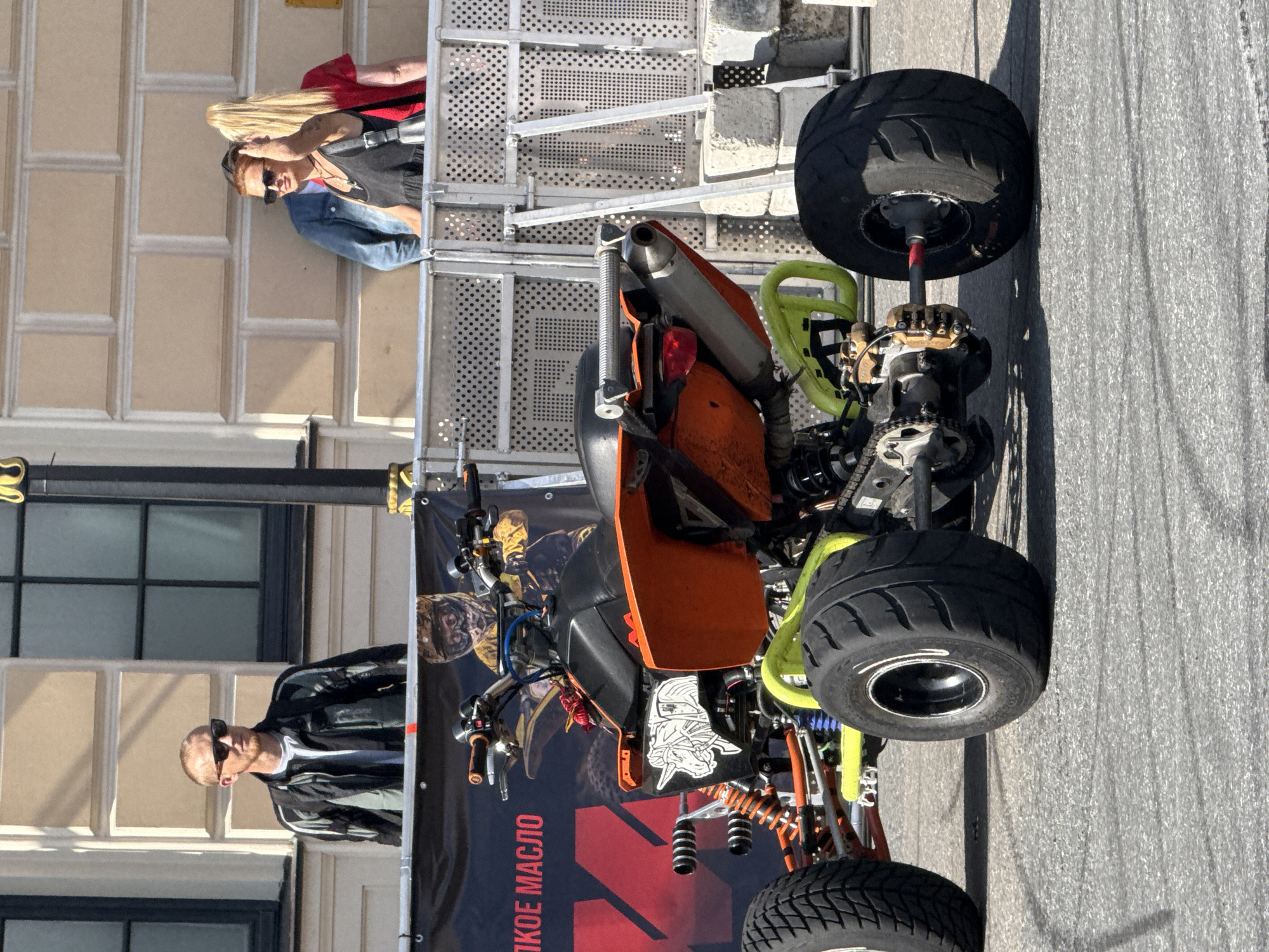 In Saint Petersburg, Russia, on a sunny day, a brightly colored orange and lime green ATV sits parked on asphalt. The ATV is detailed with a unicorn sticker on its side and has large, wide tires. Behind the ATV, a black banner with red lettering and a graphic of a person in a helmet is visible. Spectators are present, including a man with short hair and sunglasses wearing a black and gray jacket, and two women with sunglasses; one in a denim jacket and the other in a red shirt. They are standing behind a metal barrier, suggesting an event or exhibition is taking place. The background shows a building with a light-colored facade and windows, characteristic of St. Petersburg's architecture, with a street lamp on the right side.