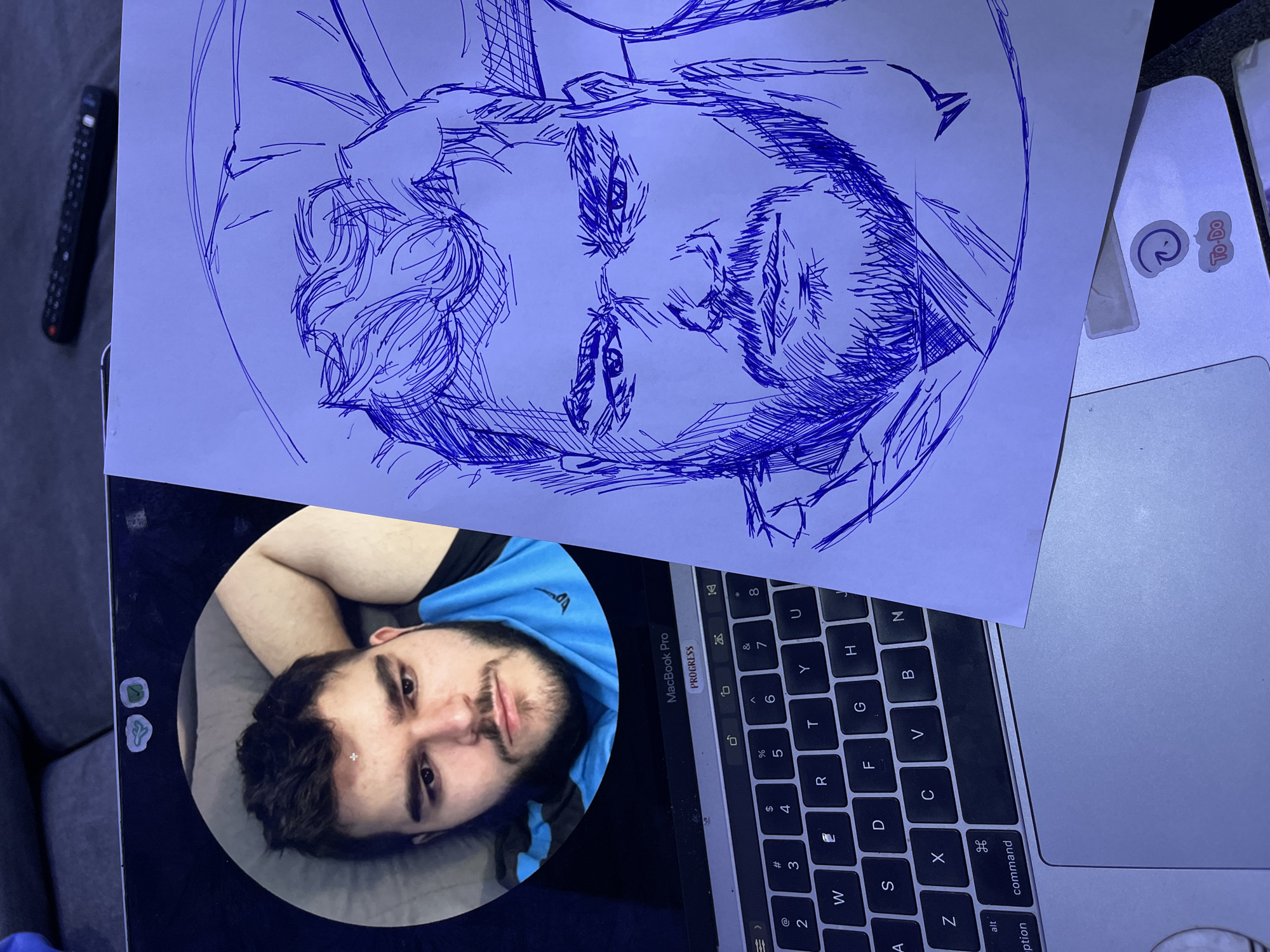 The image shows a man lying down, with his face partially visible in a circular frame on a laptop. He has dark hair and a beard, and is wearing a blue hooded shirt. A sketch of a person's face, drawn with blue ink on paper, is placed on top of the laptop keyboard. The laptop is a MacBook Pro, with visible keys and a "PROGRESS" label on a bar above the keyboard. There are stickers on the laptop, one reads "To-Do". The setting appears to be indoors, possibly a living room or office, with a gray sofa visible in the background. The overall lighting suggests it might be nighttime or a dimly lit room, with a blueish tint to the scene. The sketch is detailed, with emphasis on the eyes and hair. The man in the circular frame appears relaxed, with his eyes open and looking slightly upwards. There is no explicit indication of the time of day or weather. The location context provided is Abuja, Nigeria.