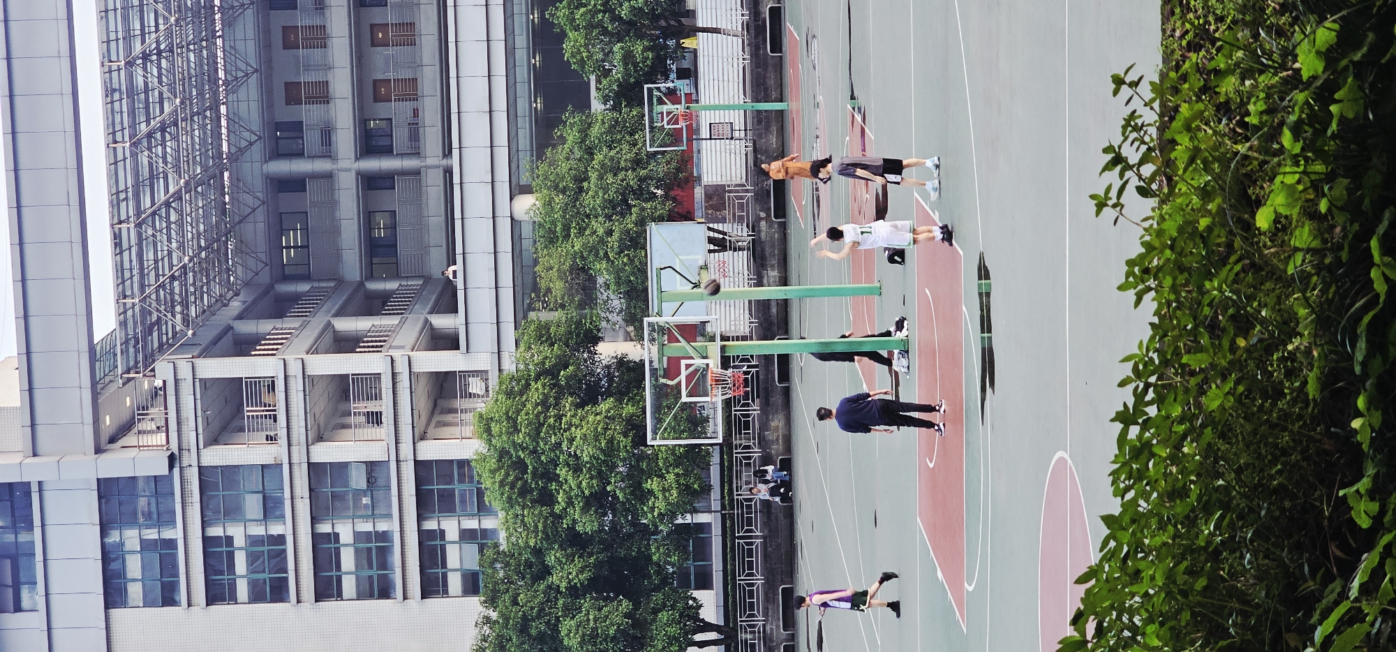 The image depicts a vertical view of a basketball court located in the Yuzhong District of China. Several individuals are present on the court, some appearing to be engaged in a basketball game. A tall building borders the court, and green trees and bushes surround the court. A basketball is airborne near one of the hoops. There is a group of people sitting in a covered seating area.