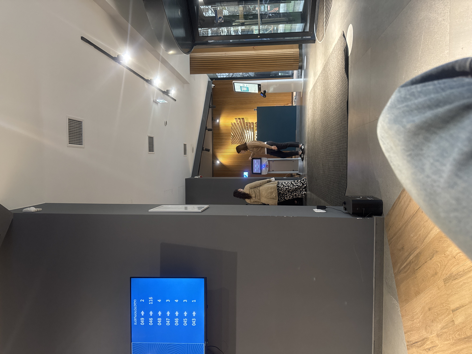 The image captures the interior of what appears to be a governmental or public service building, possibly located in Tbilisi, Georgia, given the Georgian script on the information display.
The scene is modern and sleek, with a high ceiling and contemporary lighting fixtures. On the left, a large television screen displays a queueing system with the heading in Georgian script and columns showing call numbers and corresponding counters. People are waiting, with two individuals standing near a reception or information desk towards the back of the space. One person is wearing a beige jacket and a patterned skirt, while another is wearing a brown jacket with black pants. They seem to be interacting with an automated kiosk or waiting for service.
The environment includes various modern elements like a wooden-paneled wall and a black carpet. In the foreground, a section of what seems to be a person's leg and jeans is visible, indicating the perspective of the photographer. A black rectangular object is next to the leg.