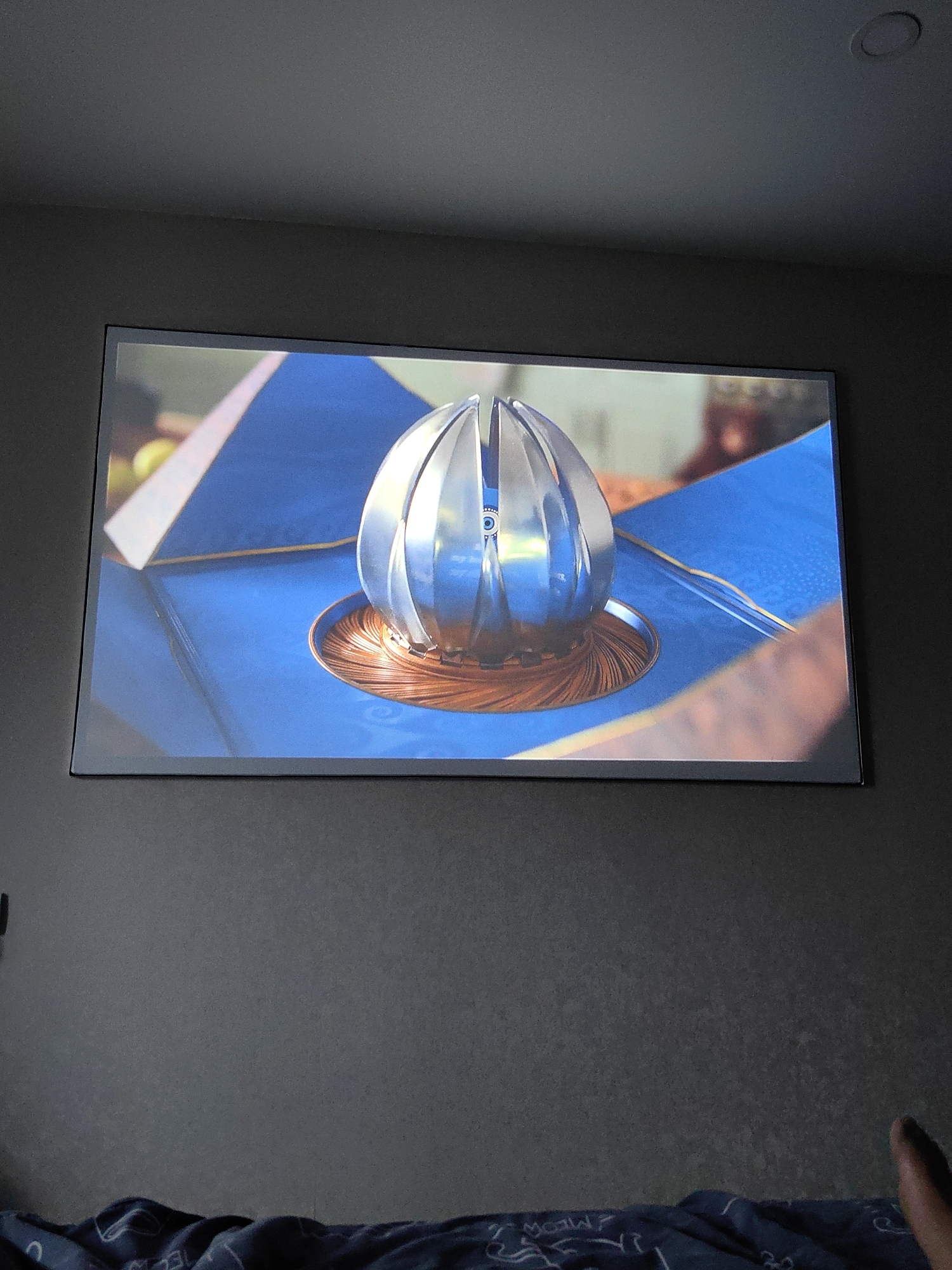 A projection screen displays an image of a metallic, spherical object with petal-like segments, situated on a circular, coppery base. The object is placed on a blue, geometric surface, with hints of other objects and colors in the background. The projection is wall-mounted in a room with dark gray walls and a ceiling with a recessed light fixture. In the foreground, a dark blue blanket with a white pattern is visible, and a person's foot is present in the lower right corner. The scene is an indoor living space.
