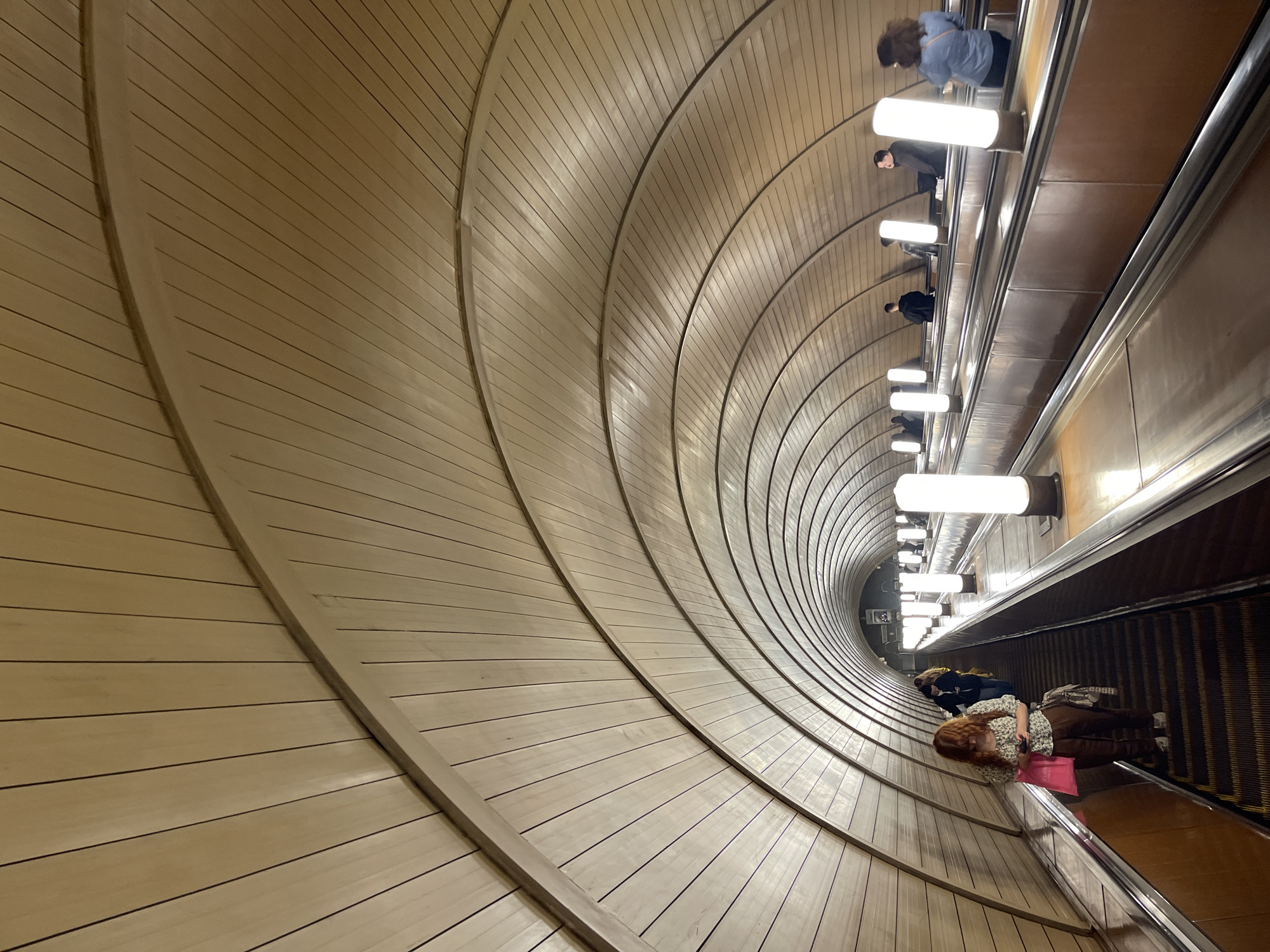 The image depicts an escalator in the Khimki metro station in Russia. The escalator ascends downwards into the station. The walls of the tunnel are constructed of light-colored wood panels arranged in concentric rings. There are people riding the escalator. Lights line the side of the escalator. One person is at the bottom of the escalator holding a pink bag.