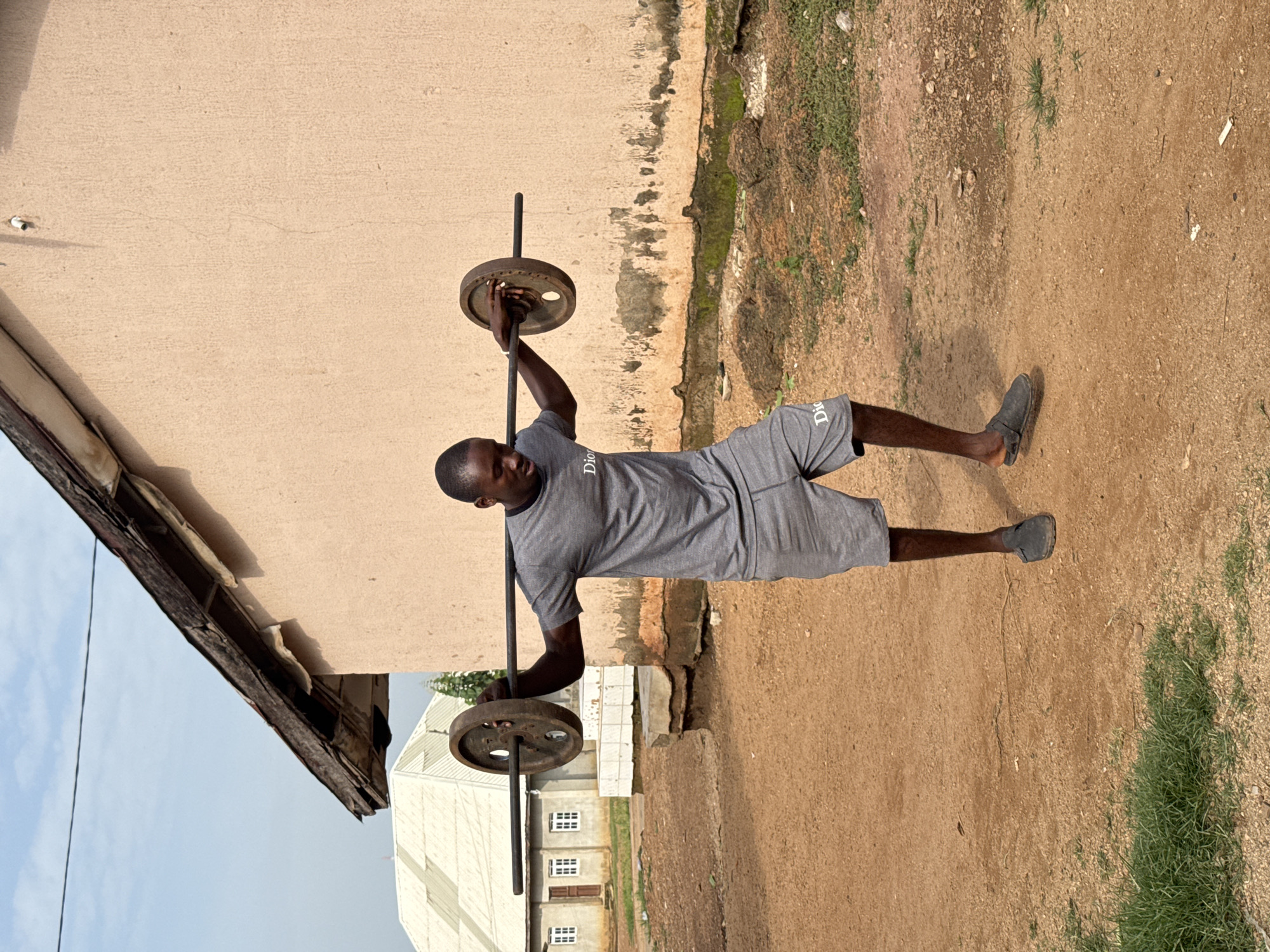 A young Black man is shown outdoors in Akwanga, Nigeria, exercising with a barbell. He is wearing a grey t-shirt with "Dior" printed on it and matching grey shorts, and dark grey slip-on shoes. The barbell, with two heavy circular weights attached to each end, rests across his shoulders. He is looking down at the barbell with a focused expression. The ground is a mix of dirt and sparse grass. In the background, a light-colored, stucco-walled building with a weathered, corrugated metal roof stands under a partly cloudy sky. To the left of this building, another structure with a white, angular roof is visible. The scene appears to be in a somewhat neglected outdoor area, possibly a compound or training ground.