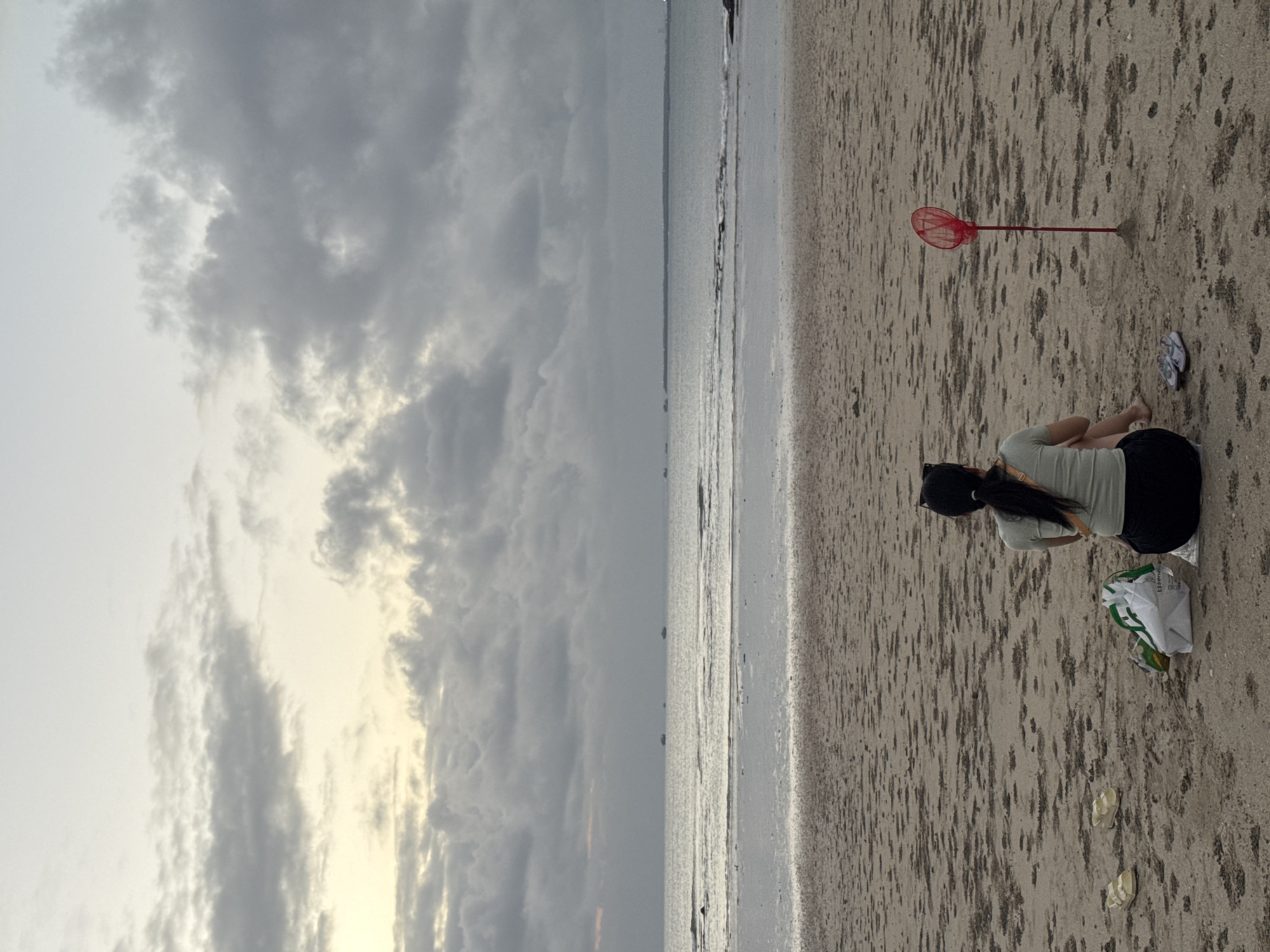 A rear view of a person is captured from the waist up, sitting on a sandy beach in Kuta Selatan, Indonesia. The individual is wearing a light green t-shirt and black shorts, with their dark hair tied back. Beside them, on the sand, are a pair of light-colored sandals and a white shopping bag with green accents. A bright red butterfly net, planted in the sand, stands a short distance away. The background reveals a calm ocean stretching to the horizon, with several small boats visible in the distance. Above the water, the sky is filled with dramatic, grey clouds, suggesting an overcast day or approaching weather. The beach itself is sandy with some scattered indentations, possibly from the receding tide.