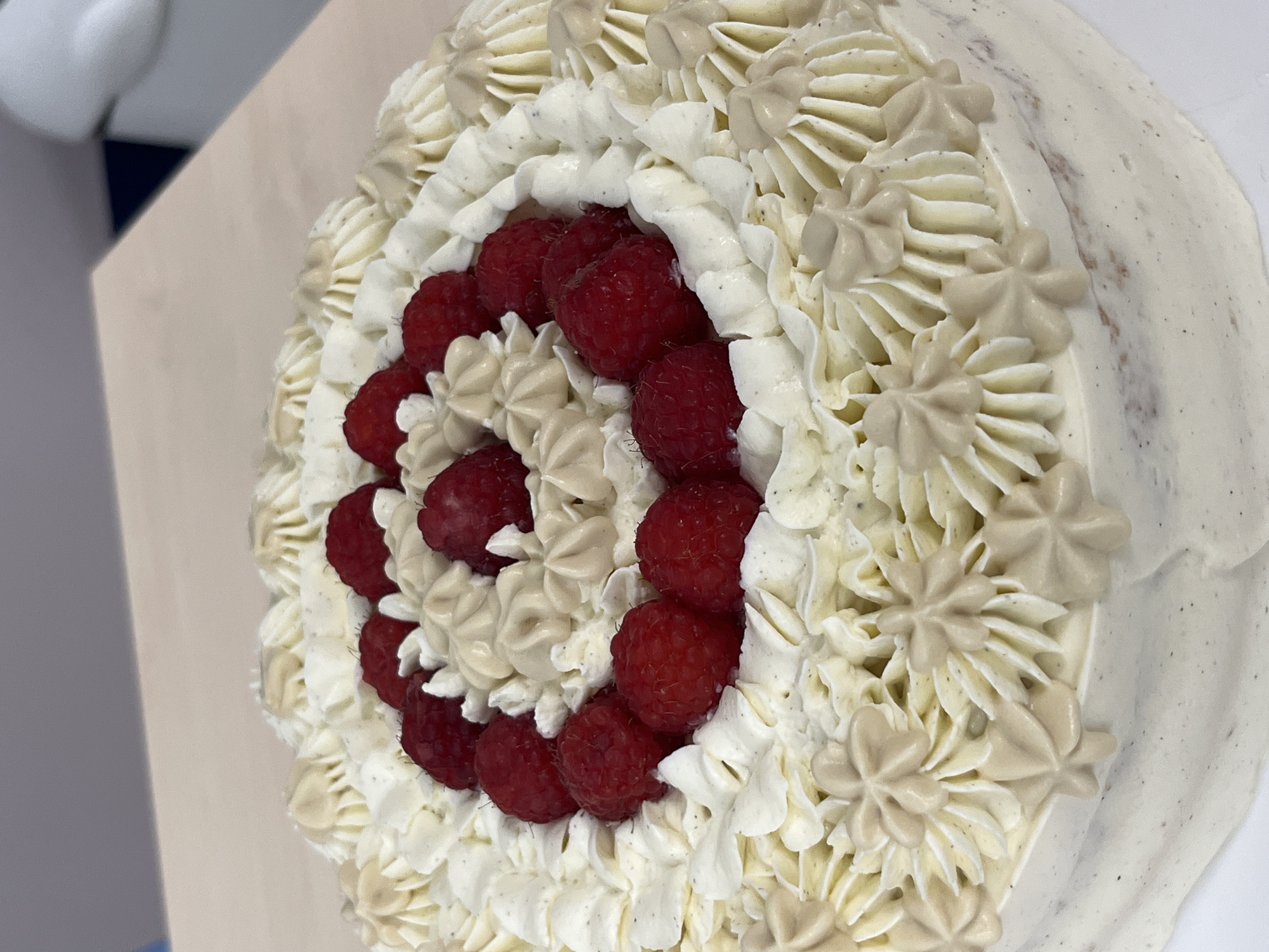 This is a close-up, overhead shot of a white frosted cake decorated with swirls of white and light brown frosting and topped with fresh raspberries. The cake sits on a light-colored, smooth surface, likely a table or countertop. In the background, a darker gray and blue surface is visible, suggesting an indoor environment. The lighting is bright, highlighting the texture and details of the frosting and fruit. No specific location in Plovdiv, Bulgaria, can be determined from the image.