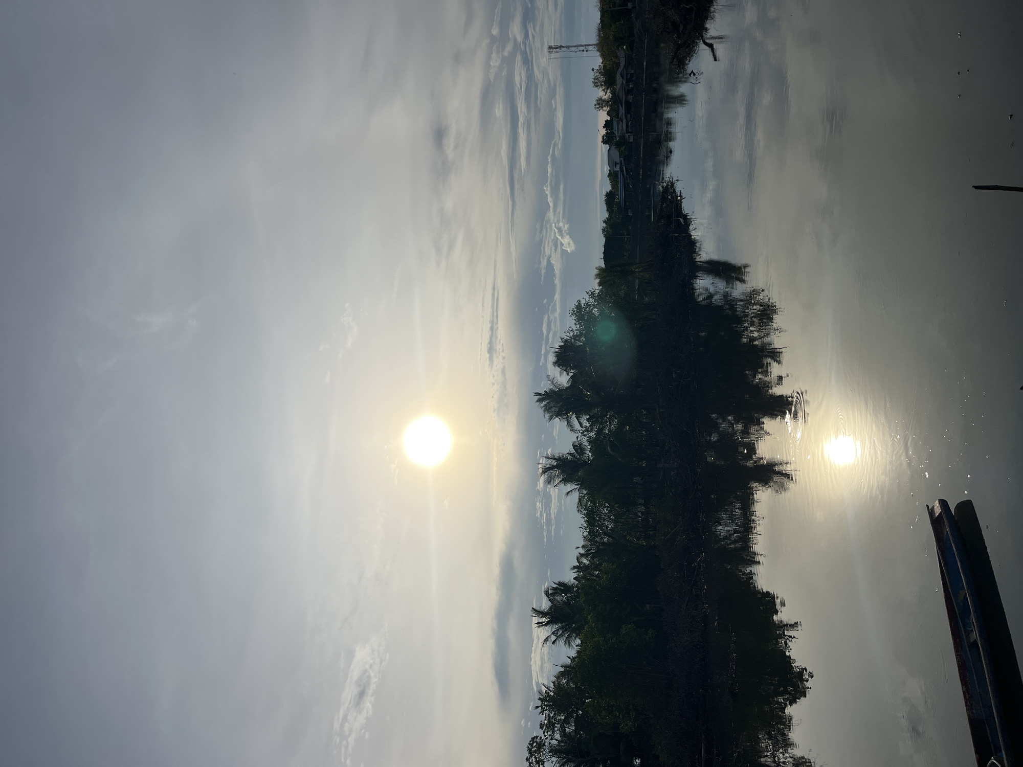 A sunlit scene depicts a body of water reflecting the sky and a line of trees, likely located in Lhokseumawe, Indonesia. The sun appears as a bright orb in the sky, and its reflection is visible on the water's surface. Silhouetted trees line the edge of the water, and what appears to be a boat is visible near the bottom right of the frame. In the background, structures, including a communications tower, are visible. The sky is overcast.