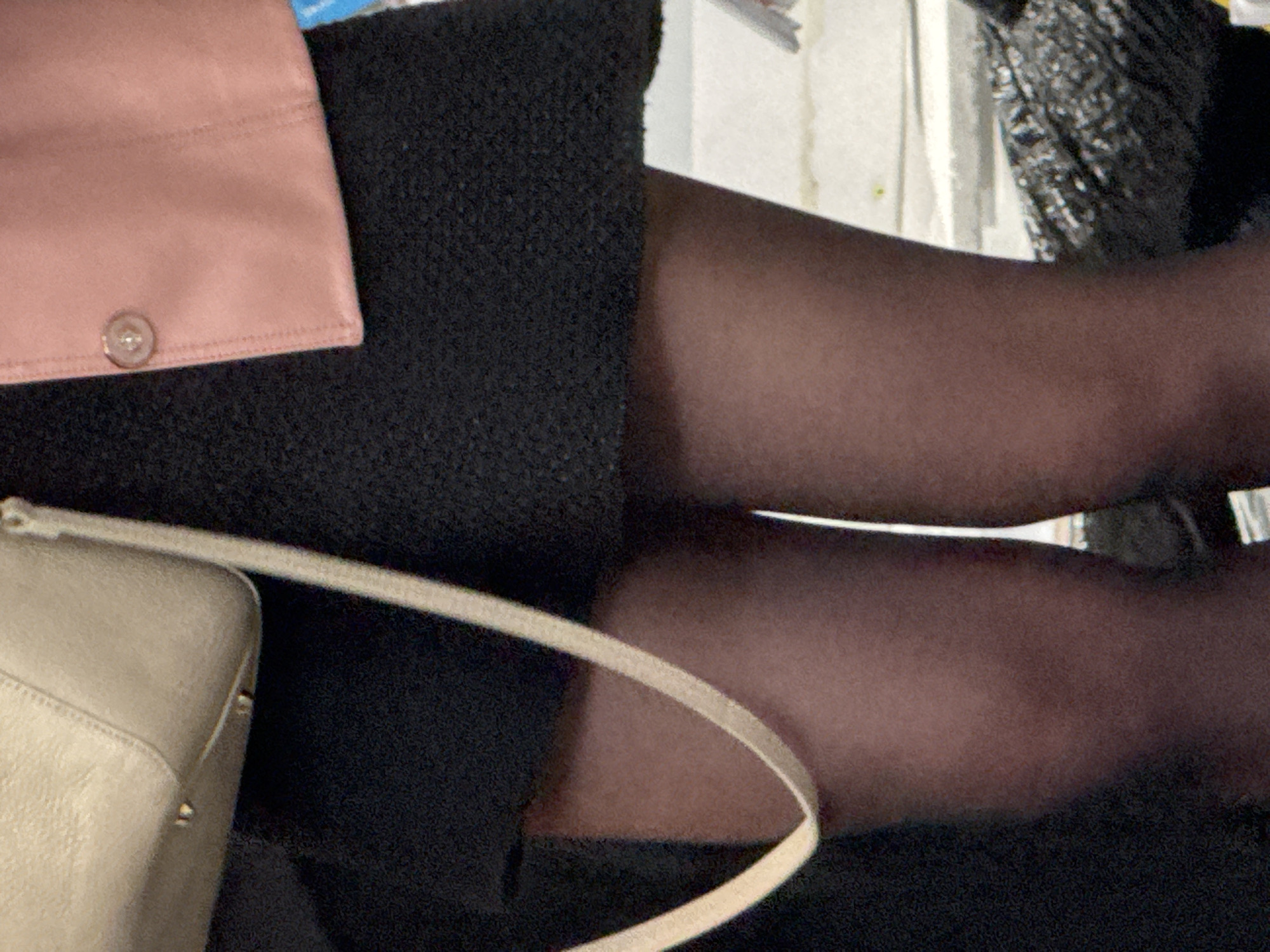 The image shows a person's legs clad in dark stockings, crossed. The person is wearing a black dress or skirt. Adjacent to the dress/skirt is a light-colored handbag with a strap and a pink garment with a button. The setting appears to be indoors. There is no indication that it is taken in Sofia, Bulgaria.