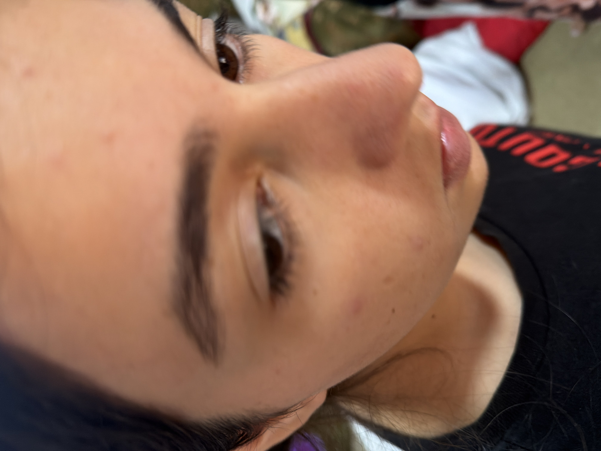 The image is a close-up, low-angle shot of a person's face, focusing on their nose, mouth, and right eye. The person has brown eyes and appears to be looking downwards. There are a few small blemishes on their skin. They are wearing a black t-shirt with red lettering. The background is out of focus, showing hints of clothing and possibly furniture. The overall lighting suggests an indoor setting. There is no information in the image to indicate its location in Köhnə Quşçu, Azerbaijan.