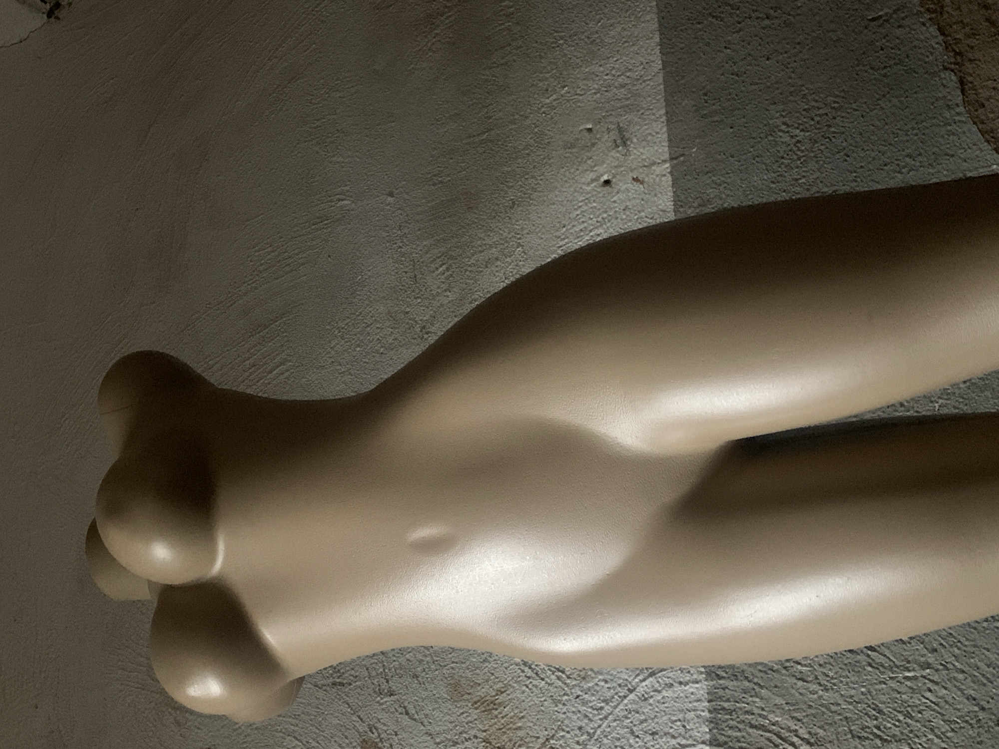 The media file displays a close-up, angled shot of a light beige, mannequin torso, possibly a partial female form, lying on a textured, gray surface. The background appears to be a rough, concrete-like wall with some subtle variations in color and texture, suggesting an indoor or sheltered outdoor location. Shadows are cast across the surface and the mannequin, indicating a light source, likely from the upper left, and implying it is daytime. The mannequin's form is smooth and rounded, with subtle indentations suggesting the suggestion of anatomical features like ribs or a navel, though these are not clearly defined. The texture of the mannequin is uniform and appears to be a matte or satin finish. The overall scene is neutral and lacks any discernible activity or emotional cues. No text is visible in the image. The image's composition focuses solely on the object and its immediate surroundings, creating an abstract or minimalist aesthetic.