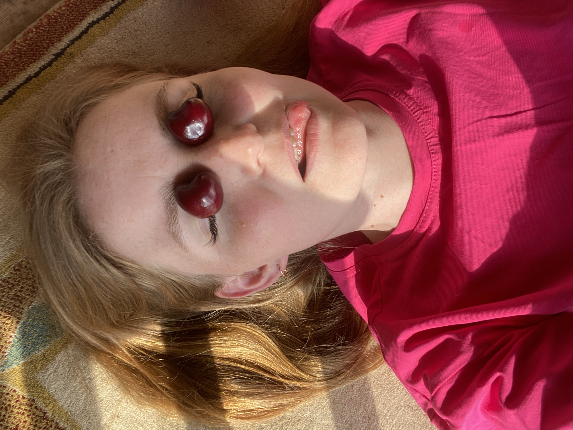 A young woman with blonde hair is lying down outdoors, possibly on a rug or blanket, as indicated by the textured surface beneath her. She is wearing a bright pink t-shirt. Two cherries are placed on her eyelids, and her mouth is slightly open, with her tongue visible. Her hair is illuminated by sunlight, creating highlights and shadows. The background appears to be outdoors, with a patterned rug and sunlight casting shadows. There is no discernible information in the image that would indicate a specific location within the city of Chaikovsky, Russia.