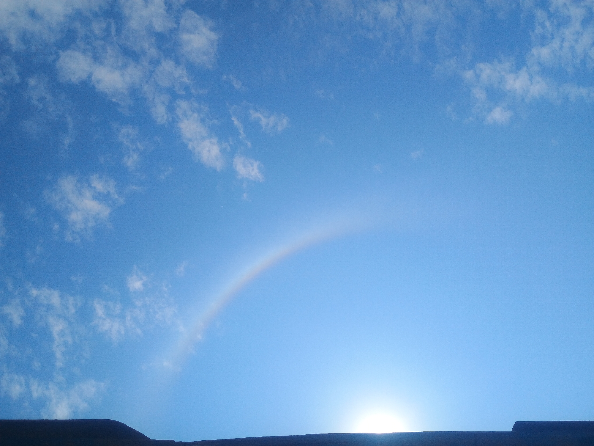 The image captures a bright blue sky with scattered white clouds, hinting at fair weather. On the left side of the frame, a dark, vertical structure, possibly a building or a portion of one, is visible, casting a shadow that encroaches into the sky. A faint, arced rainbow is present in the upper left quadrant of the sky, partially obscured by the sun's glare which is also located in the lower left of the frame. The scene appears to be an outdoor shot taken from a low angle, looking upwards. There are no people or animals visible in the image. The environment is natural, focusing on the sky and its atmospheric phenomena.