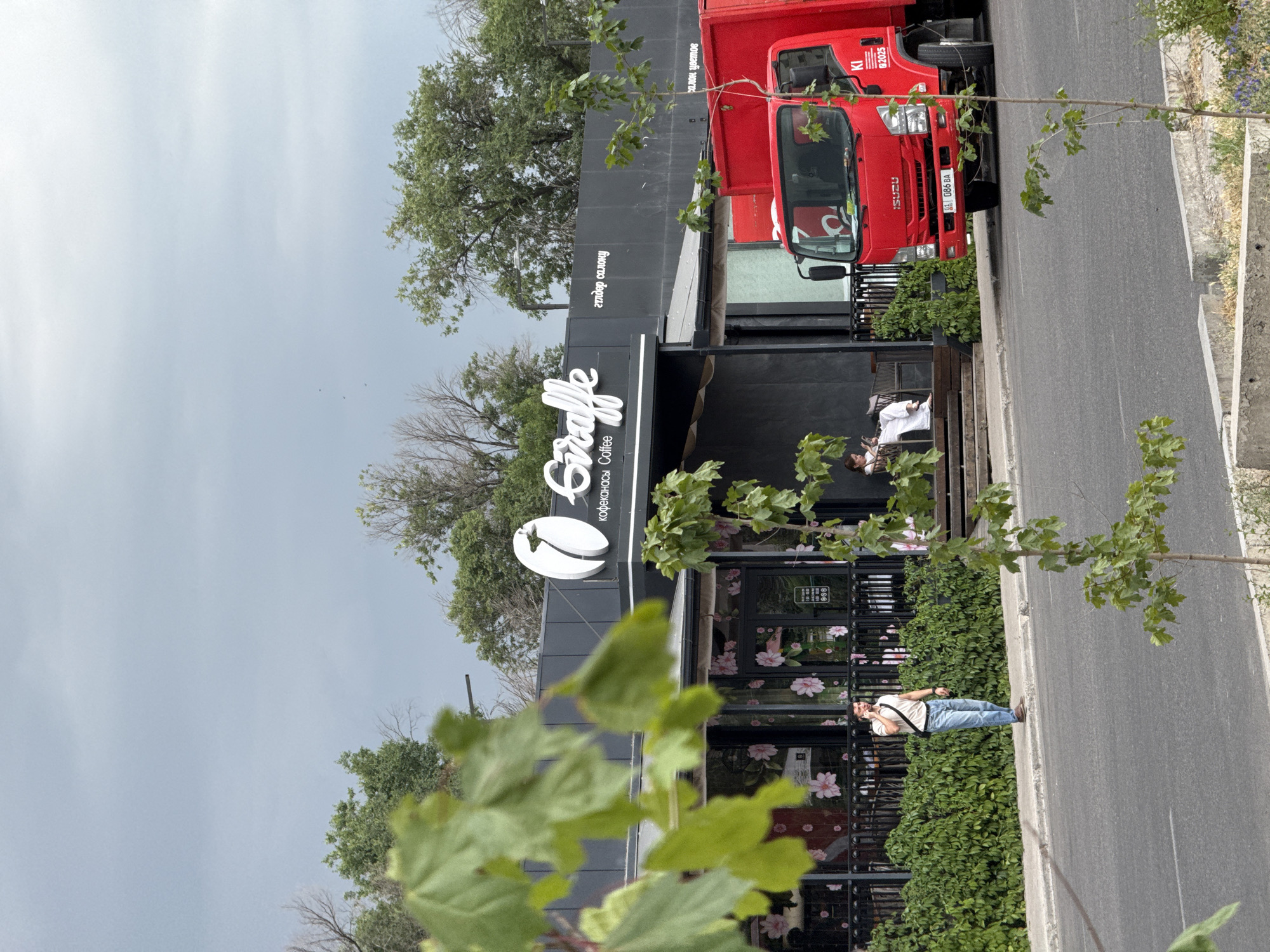 The image shows a street scene in front of a building with signage that includes the word "Giraffe" and "Coffee". A man is standing on the sidewalk, talking on his phone. He is wearing a light-colored polo shirt, blue jeans, and brown shoes. A red Isuzu truck is parked on the street next to the building. The building has large windows decorated with floral patterns and a black awning. Green ivy covers a portion of the building's facade. Trees and foliage are visible in the background, and the sky is overcast.