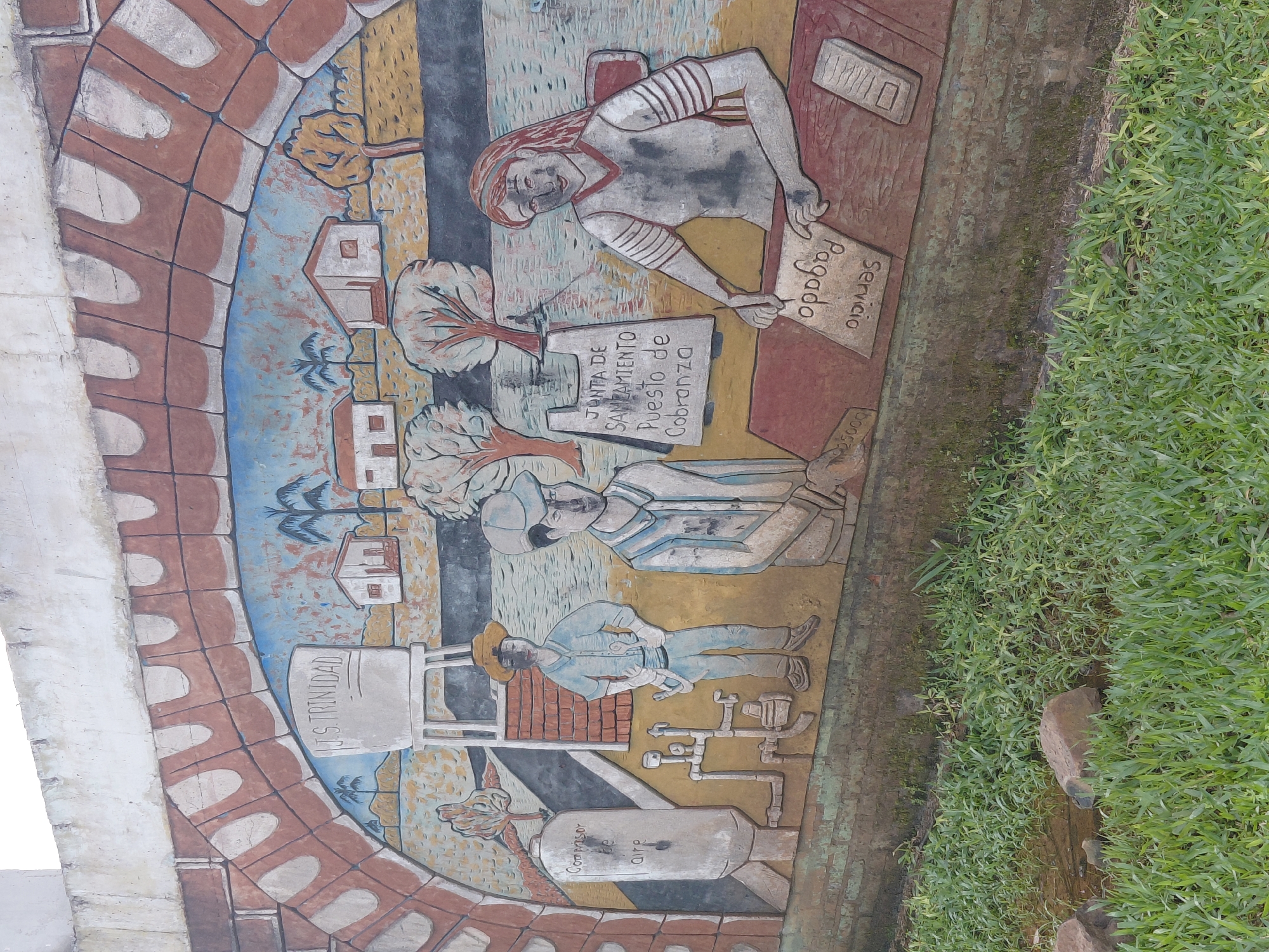 This is a photograph of a mural painted on a wall in the city of Trinidad, Paraguay. The mural depicts a rural scene with several figures. In the background, there are houses, trees, and a water tower with "J.S. TRINIDAD" written on it. In the foreground, there are two men and a woman. One man is standing and appears to be a worker, wearing a hat and holding his hands on his hips. Beside him, another man, also dressed in work clothes, stands with his hands clasped. The woman is seated behind a desk, appearing to be an administrator or cashier, with papers in front of her. One paper has "Servicio Pagado" written on it. The mural is colorful, with vibrant blues, reds, and yellows used in the painting. The wall itself is made of brick and concrete, and the mural is painted directly onto its surface. The lower portion of the wall is covered in moss and vegetation, with grass and water visible in front of it.