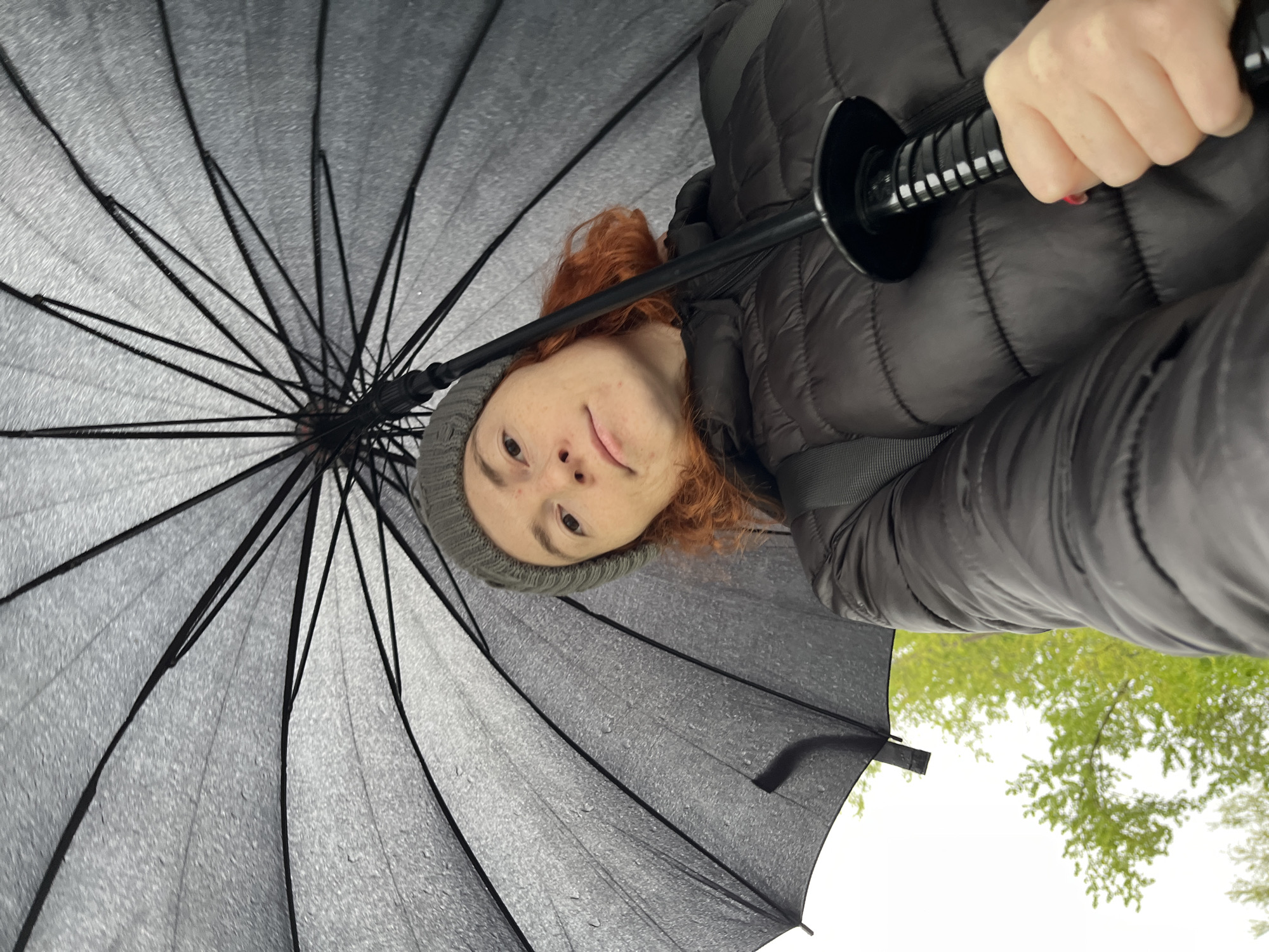 A person with reddish-brown hair and wearing a grey beanie and a dark, quilted jacket is holding open a black umbrella in Chaikovsky, Russia. The umbrella is positioned above the person's head, providing shelter, and the view is directed upwards through the umbrella. A glimpse of trees and the sky is visible outside the perimeter of the umbrella. The umbrella is damp, suggesting that it is raining or has recently rained. The person is looking down at the camera.