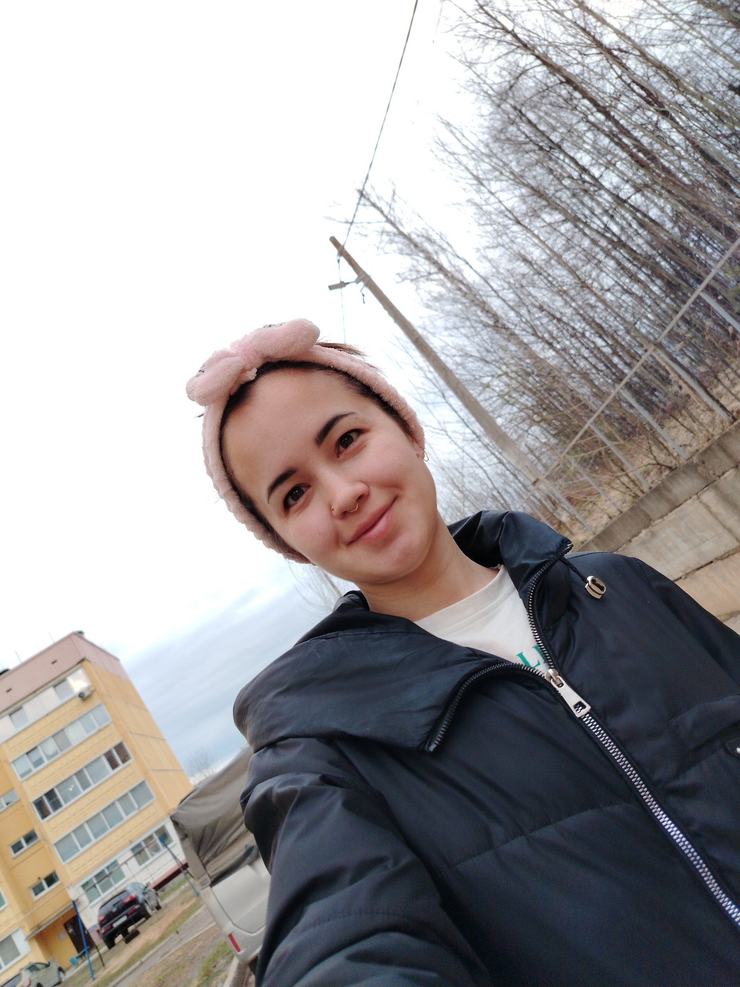 A young woman is featured in a close-up outdoor selfie, likely taken in Noviy, Russia. She has dark hair pulled back and wears a distinctive pink, fluffy headband shaped like a bow. Her expression is a gentle smile, and she looks directly at the camera. A gold-colored ring is visible in her left nostril. She is dressed in a black puffer jacket with a prominent silver zipper, and a white t-shirt underneath, which features some indistinct green text. A gold heart-shaped pin adorns the lapel of her jacket.
The background on the left shows a multi-story yellow apartment building with white-framed windows, and several cars are parked on the ground in front of it. On the right, a cluster of bare, leafless trees with thin branches stands against a bright, overcast sky. A utility pole or fence post and a simple metal fence are also visible in this area. The cloudy sky suggests it is daytime, possibly during late autumn or winter due to the barren trees.