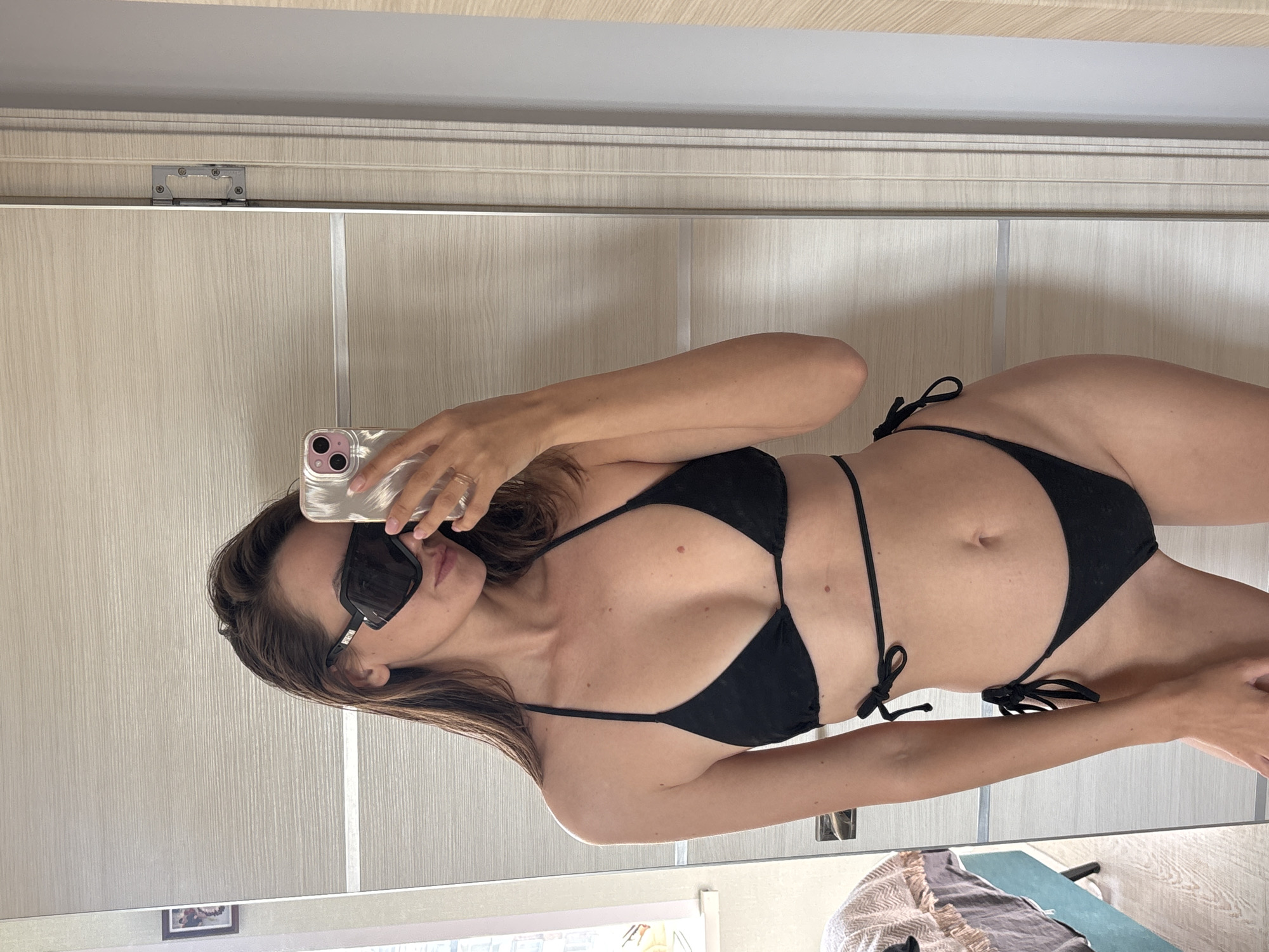 The image is a full-body mirror selfie of a woman, taken indoors in what is identified as Krasnoyarsk, Russia. The original photo is oriented sideways, but the subject is depicted as standing upright. The woman has long, dark brown hair and is wearing dark rectangular sunglasses and a black two-piece string bikini with tie fastenings. She appears to be in her 20s or 30s with a slender build. She holds a smartphone, encased in a light pink or clear cover, in her left hand to take the picture; a ring is visible on her left ring finger. Her expression is partially obscured by the sunglasses, but her lips are slightly parted, conveying a relaxed and confident demeanor.
The reflection in the mirror reveals an indoor setting, likely a bedroom or dressing area. The background primarily consists of a light-colored wall or wardrobe with a subtle vertical wood-grain pattern. A silver hinge is visible on a panel or door near the top left of the reflection. The ceiling above and the floor below are both light-toned. In the bottom right corner of the reflection, a patterned blanket or throw is draped over a piece of furniture, with a glimpse of blue fabric next to it. A small, indistinct framed picture can be seen reflected on a wall in the lower left. The lighting is bright and even, indicative of daytime. No visible text is present in the image. The scene suggests a casual personal moment.