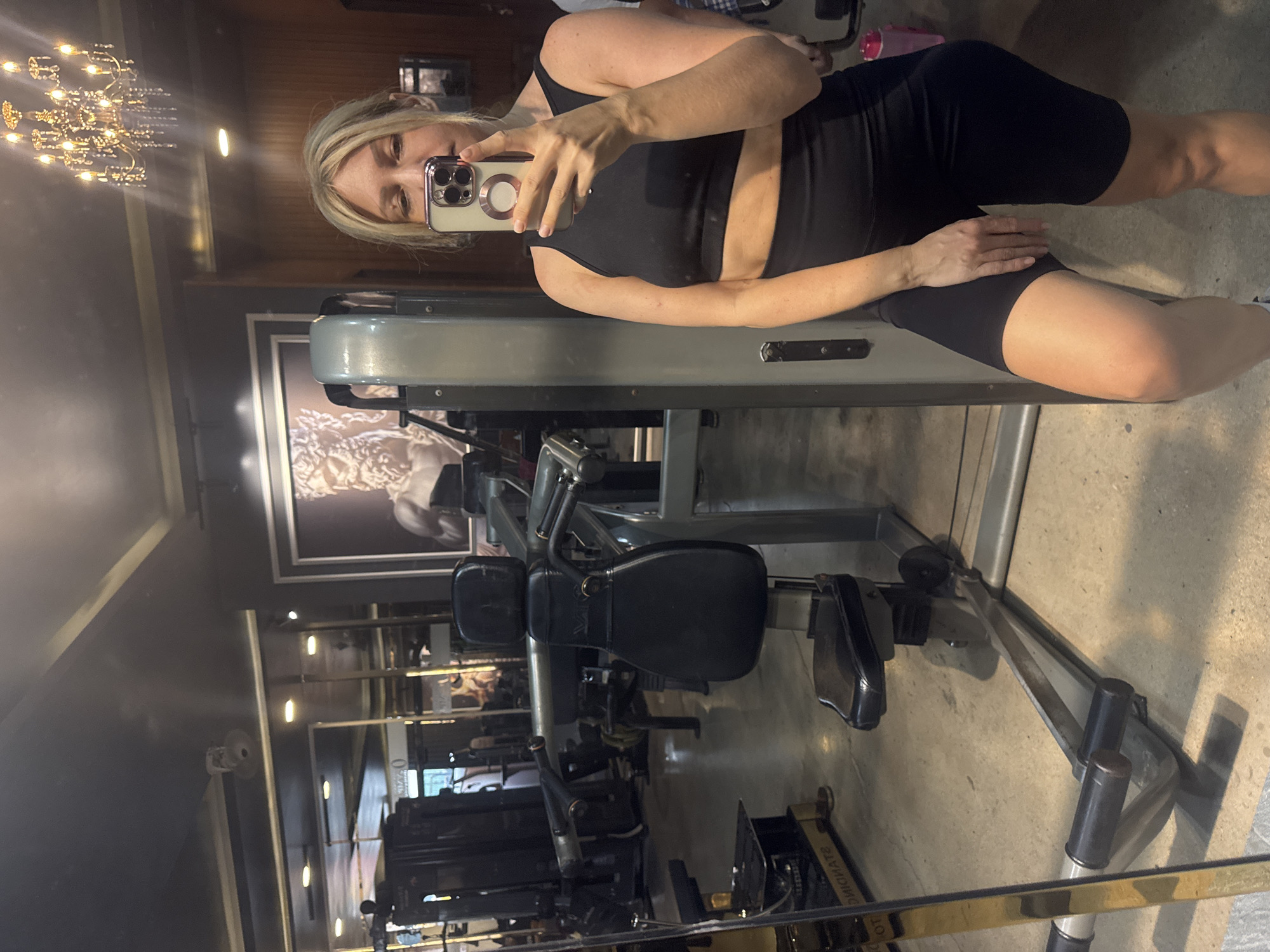 A woman stands in a gym in Nha Trang Ward, Vietnam, taking a selfie in a mirror. She is wearing a black sports bra and black biker shorts. The gym has various weight machines and a large framed picture on the wall. A chandelier hangs from the ceiling.