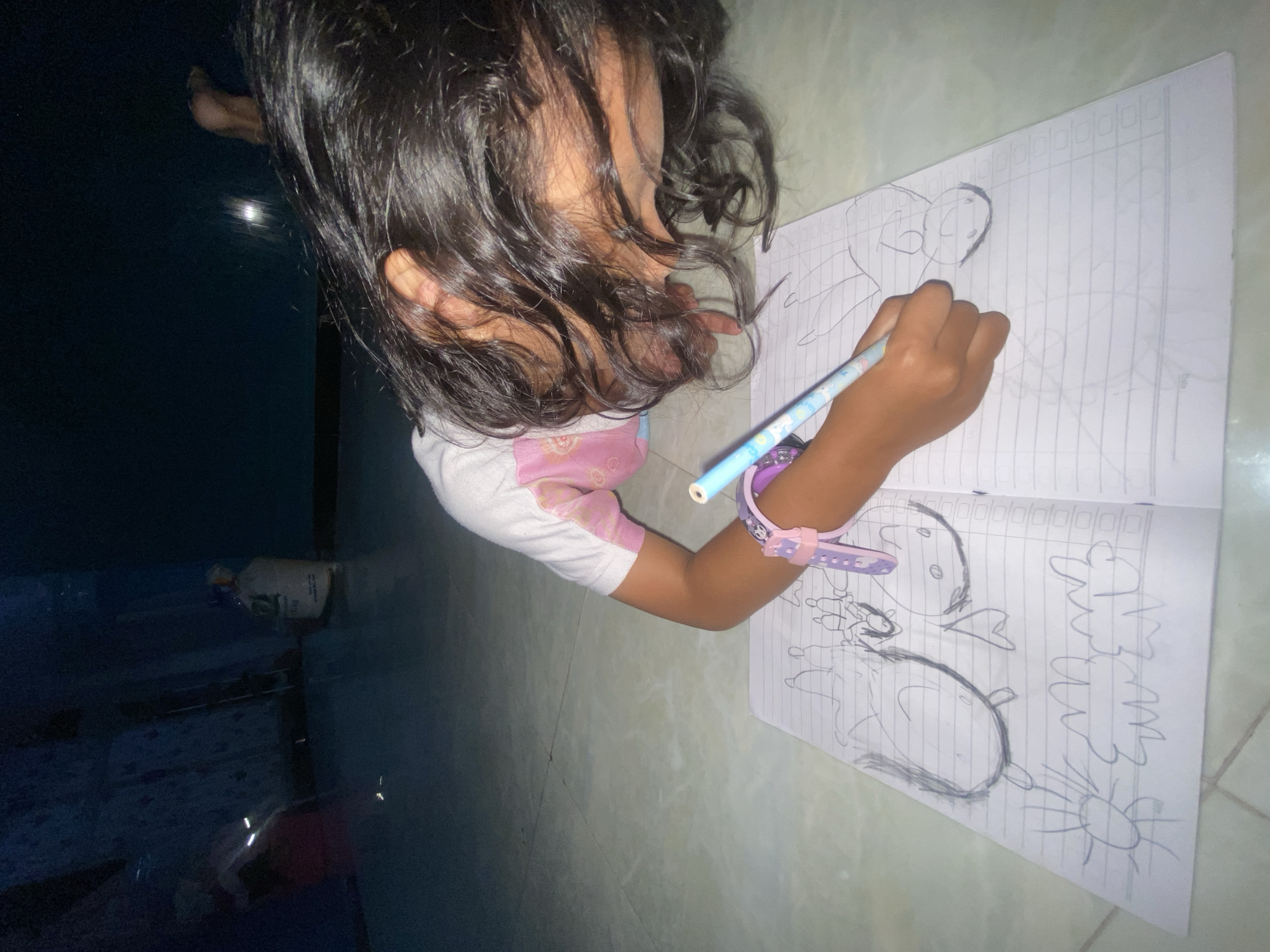 A young girl is lying on her stomach on a tiled floor, drawing in a notebook. She is wearing a pink and white t-shirt and has dark, long hair. She is holding a blue pencil in her right hand and appears to be concentrating on her drawing. On her left wrist, she is wearing a purple and pink digital watch. The notebook is open to a page filled with simple line drawings of animals, shapes, and what appear to be people. The background is dark, with some objects visible in the dim light, including a white bag on the right side of the frame. The overall scene suggests a moment of creative activity within a home environment, likely in Gelam, Indonesia, given the context.