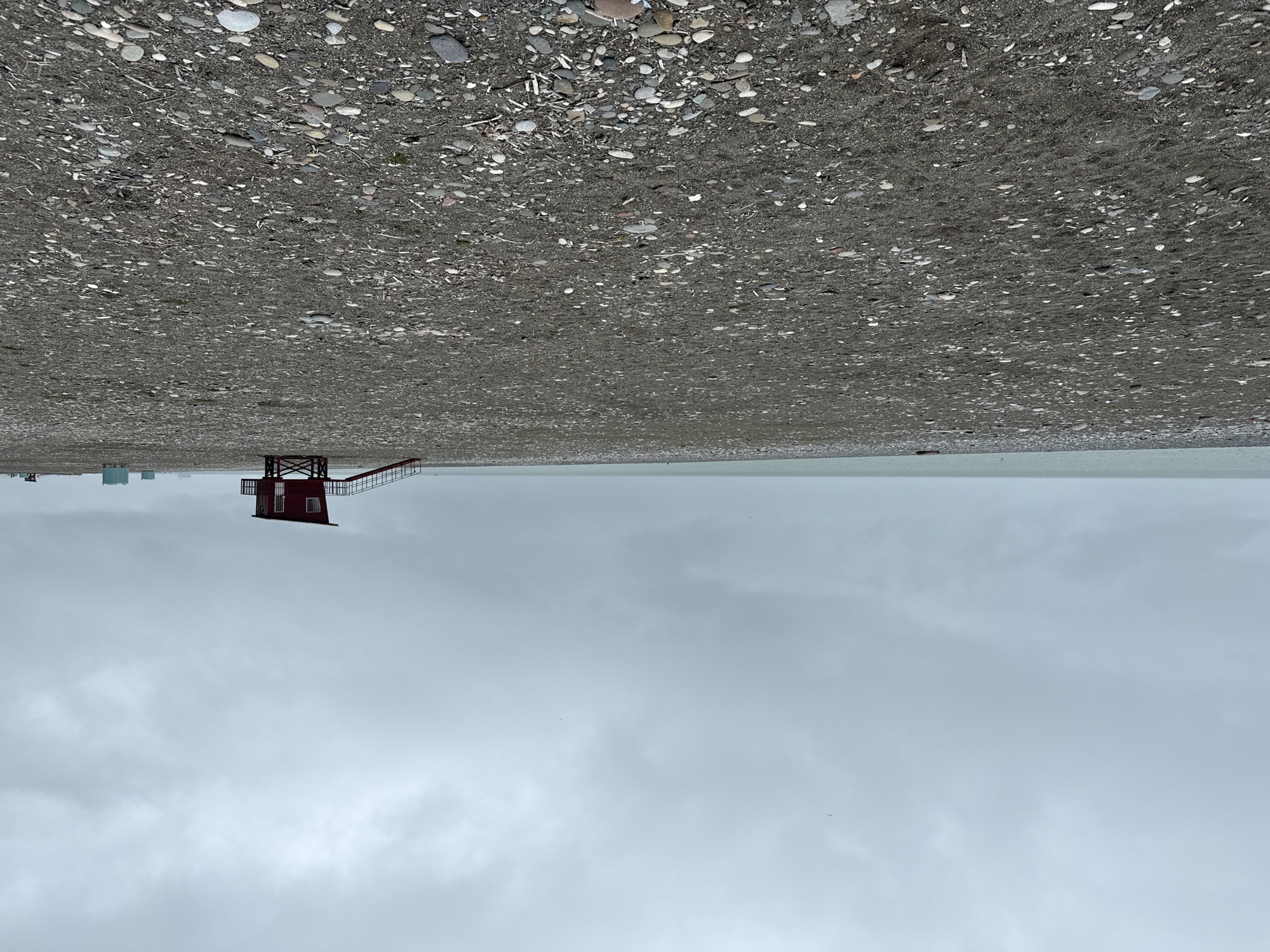 The image depicts a beach scene in Batumi, Georgia, with an overcast sky dominating the view. The photograph is taken from a low perspective and inverted. A red lifeguard tower is positioned near the center, appearing to be stationed on the shoreline where the beach transitions into the sea. The beach itself is composed of pebbles and stones, extending towards the top of the image. In the distance, some indistinct buildings are visible on the horizon near the left side of the frame. The sea appears calm and merges with the gray sky at the horizon, creating a blurred boundary between the two.