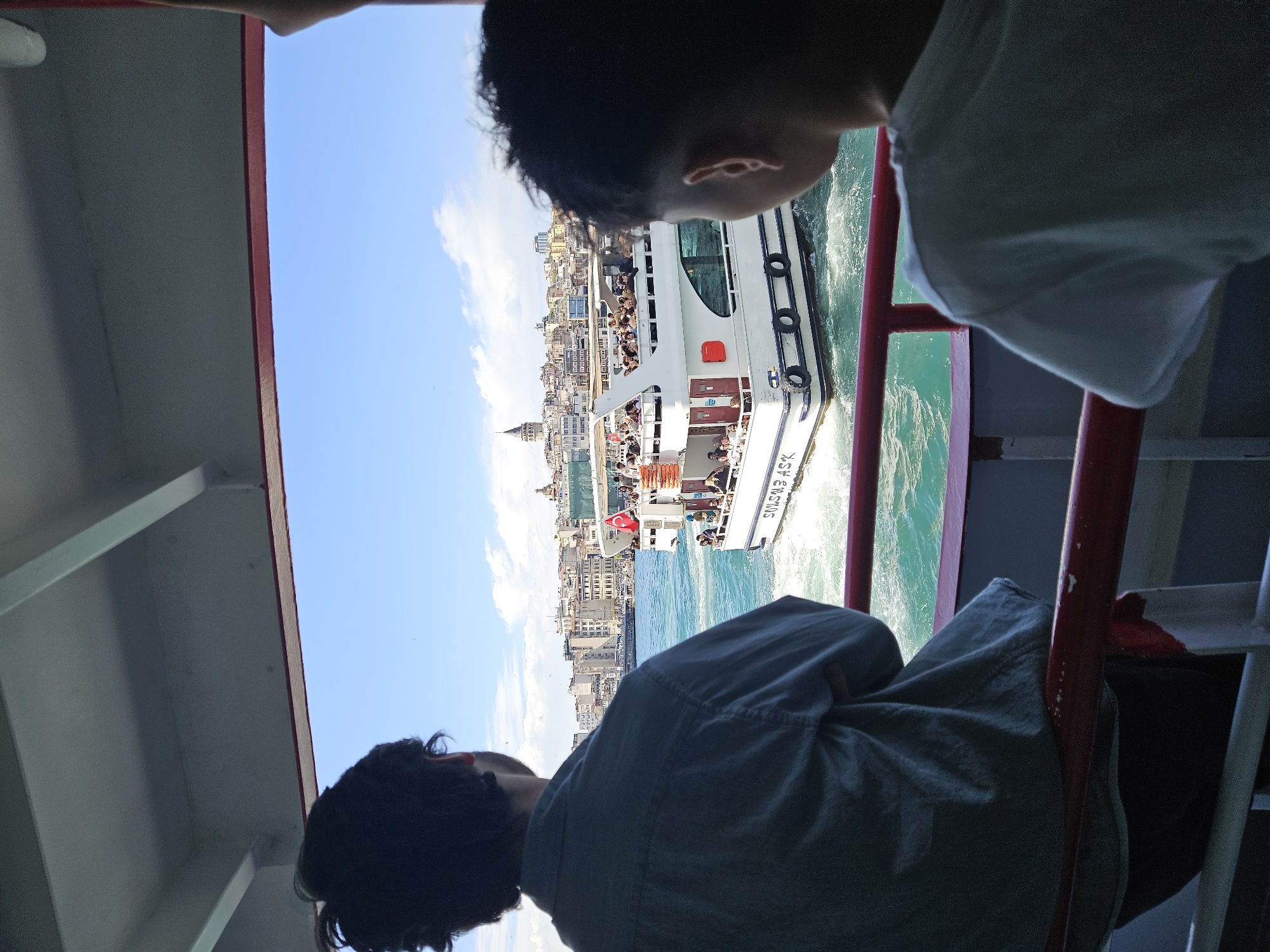 A photo of a man and a child on a boat looking at another boat full of people. The photo is taken from a side angle. The men have dark hair, and the child is wearing a white shirt. The boat is white with red accents and says "SEMS EMRINS." The city in the background is Beyoğlu, Turkey, which is identifiable by the Galata Tower. The sky is blue with some clouds. The water is turquoise and choppy. There are metal bars and part of the roof of the boat. The people are looking at the boat full of people.
