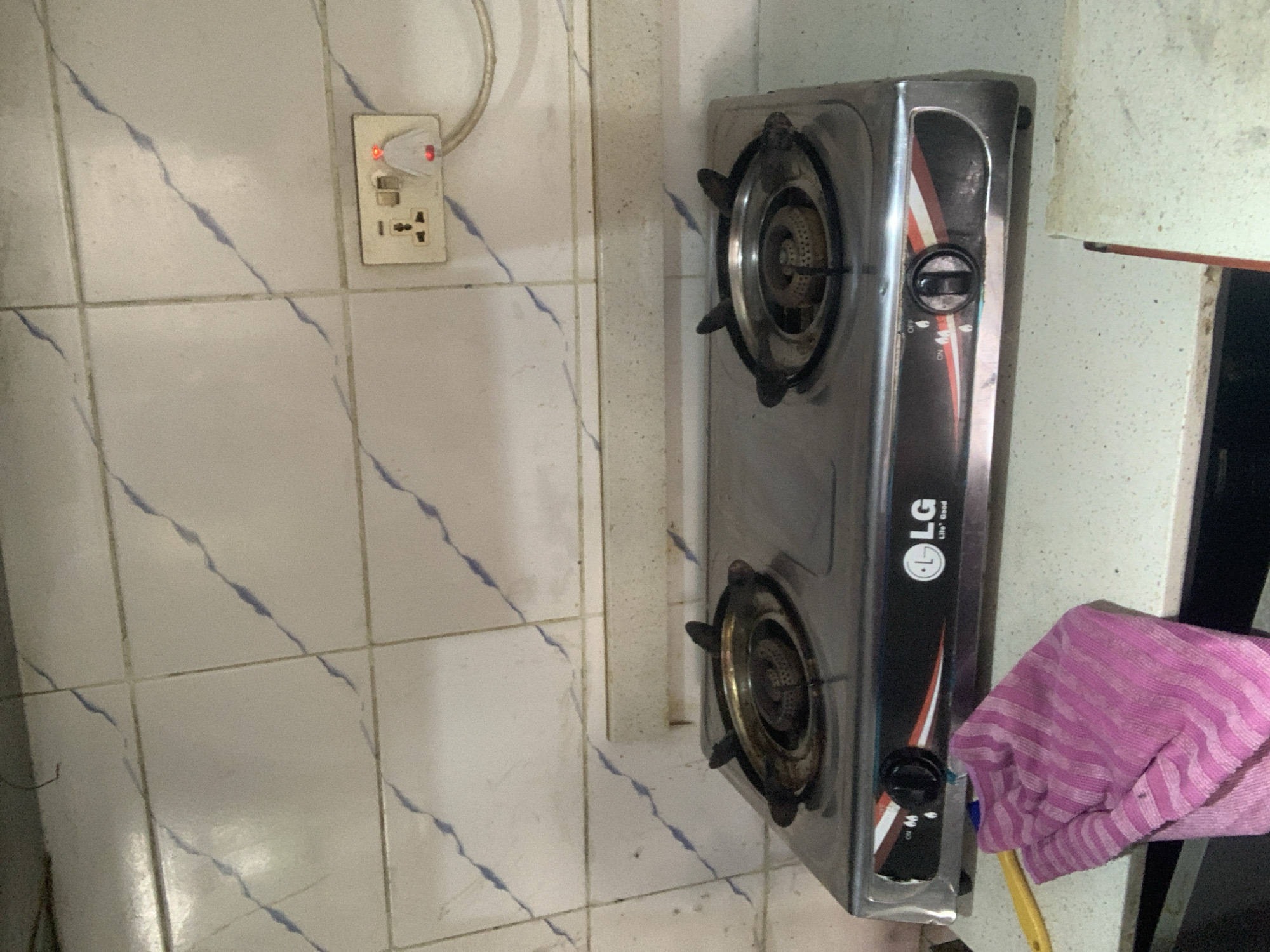 The media file shows a close-up view of a two-burner gas stove against a tiled wall. The stove is stainless steel with a black control panel that has "LG" branding and a circular logo that says "Life's Good". The control knob is black and appears to be set to "ON" as indicated by white text on the panel. To the left of the stove, a white electrical outlet is mounted on the tiled wall. A white plug with red indicator lights is inserted into the outlet. The tiles are white with thin blue diagonal lines. A pink and purple striped cloth is draped over the right side of the stove, partially obscuring the side of the appliance. The lighting suggests an indoor setting, likely a kitchen or cooking area. There is no indication of people or specific activity, other than the presence of the stove which implies cooking may take place. The scene is static, with no discernible time of day or weather conditions. The visible text includes "LG" and "Life's Good". The location context is Abuja, Nigeria.