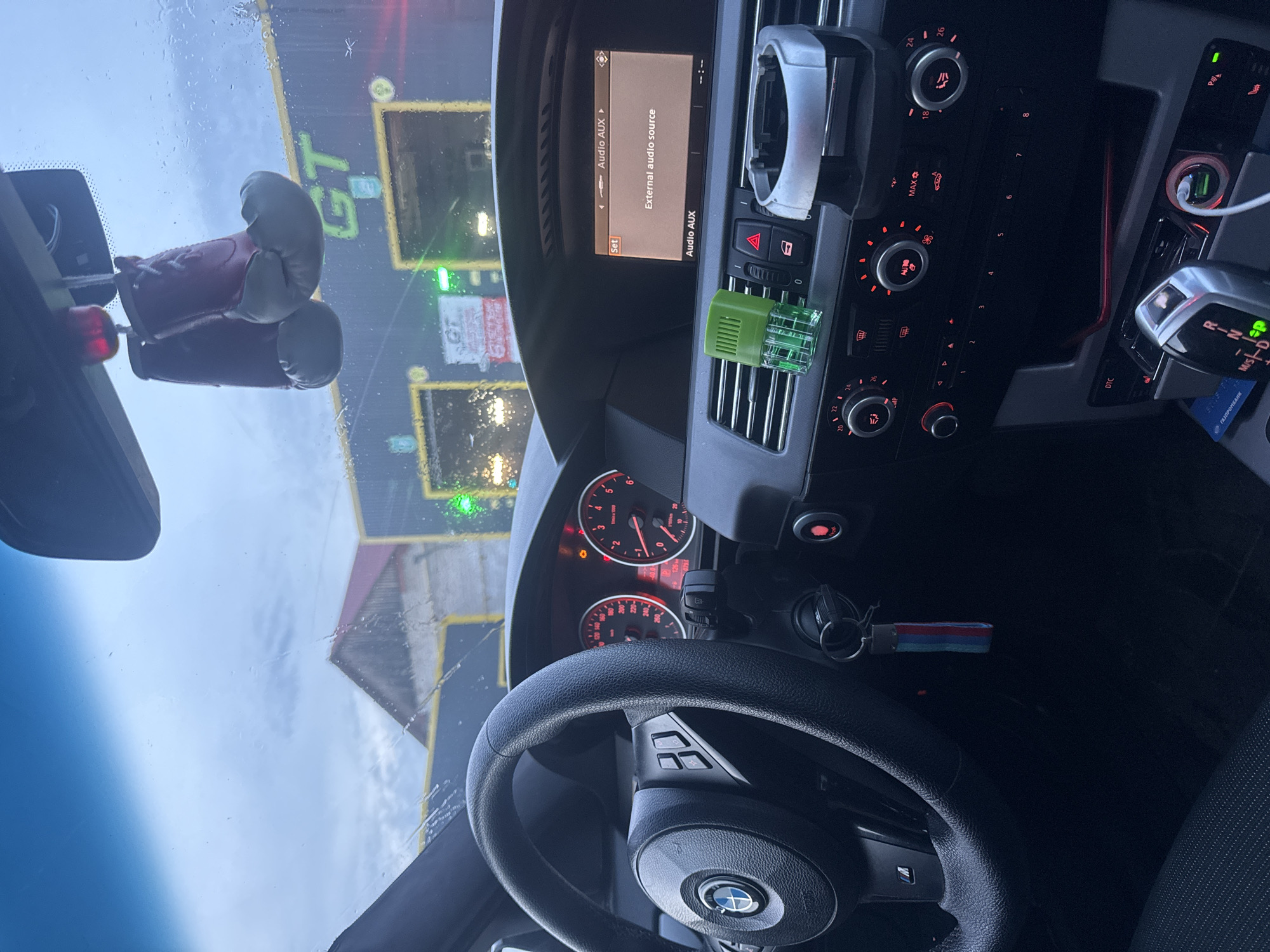 The image shows the interior of a car, likely a BMW, with the driver's perspective looking out through the windshield. Outside, a car wash facility with "GT" branding is visible. The car's dashboard is in focus, displaying the audio system interface set to "Audio AUX" and indicating "External audio source." The steering wheel and instrument panel, including the speedometer and tachometer, are also visible. A pair of miniature boxing gloves are hanging from the rearview mirror. Based on the presence of a "Газпромбанк" loyalty card, the location is likely within Russia. The presence of the "GT" branded car wash and other details indicates the location is likely in Chaikovsky.