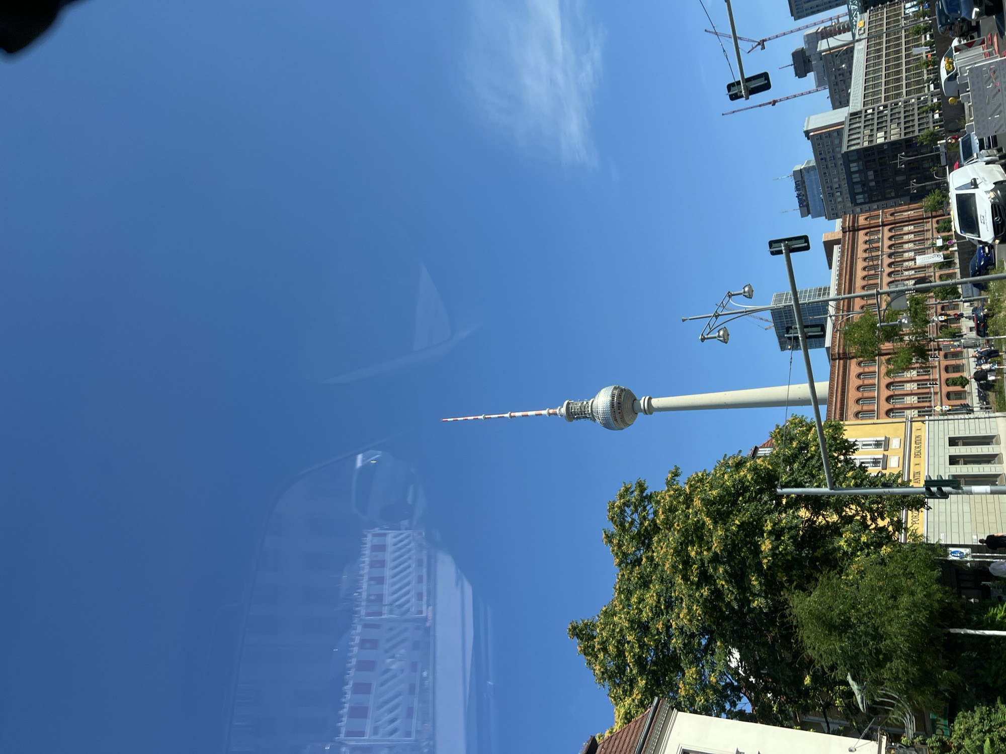 The image shows a view of Berlin, Germany, with the Fernsehturm (Television Tower) prominently displayed against a clear blue sky. The scene appears to be captured from a moving vehicle, as evidenced by reflections on the surface, likely a window.
In the foreground, several buildings line a street. One building is painted yellow with arched windows and a sign that reads "ANTIK DEKORATION". Another building to its left is white. Lush green trees, one with yellow flowers, are interspersed between the buildings and along the street. Traffic lights are visible, and several vehicles, including cars and vans, are on the road, indicating a busy urban environment. Construction barriers are also present.
The Fernsehturm stands tall in the mid-ground, its distinctive silver sphere and red and white antenna piercing the sky. Behind the tower, other modern buildings and construction cranes can be seen, suggesting ongoing development in the city.
The overall impression is a sunny day in a vibrant, active city with a mix of historic and modern architecture.