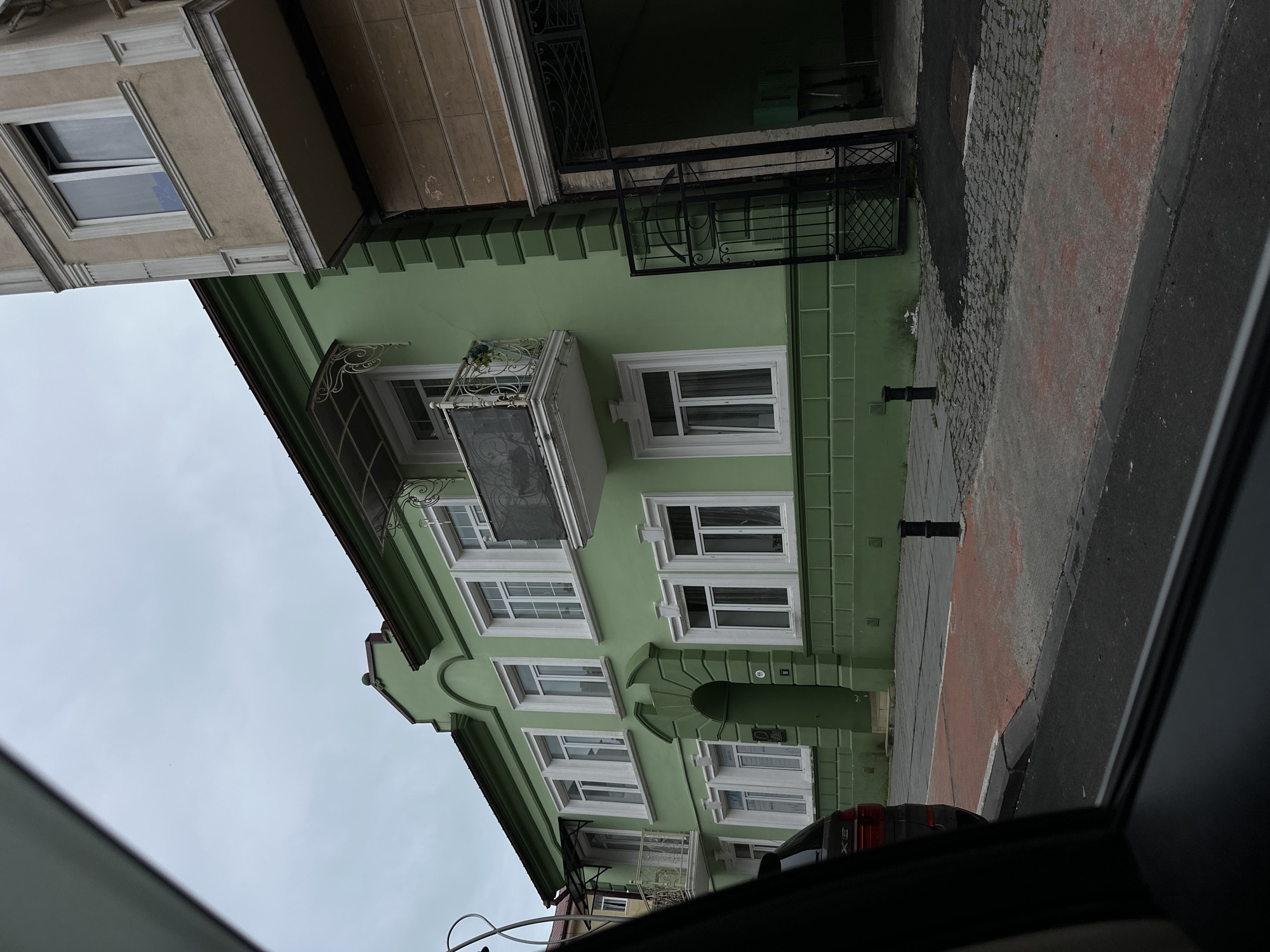 The image depicts a street scene in Batumi, Georgia, as viewed from inside a vehicle. A green-painted building with white-framed windows and architectural details occupies a significant portion of the frame. The building has a balcony with decorative ironwork. A neighboring building with a beige facade is partially visible to the left. The street is paved with cobblestones and asphalt, with a section painted red. Black bollards are placed along the sidewalk. The sky is overcast. Elements of the car's interior, such as the window frame and parts of the dashboard, are visible. There are no people visible in the scene.