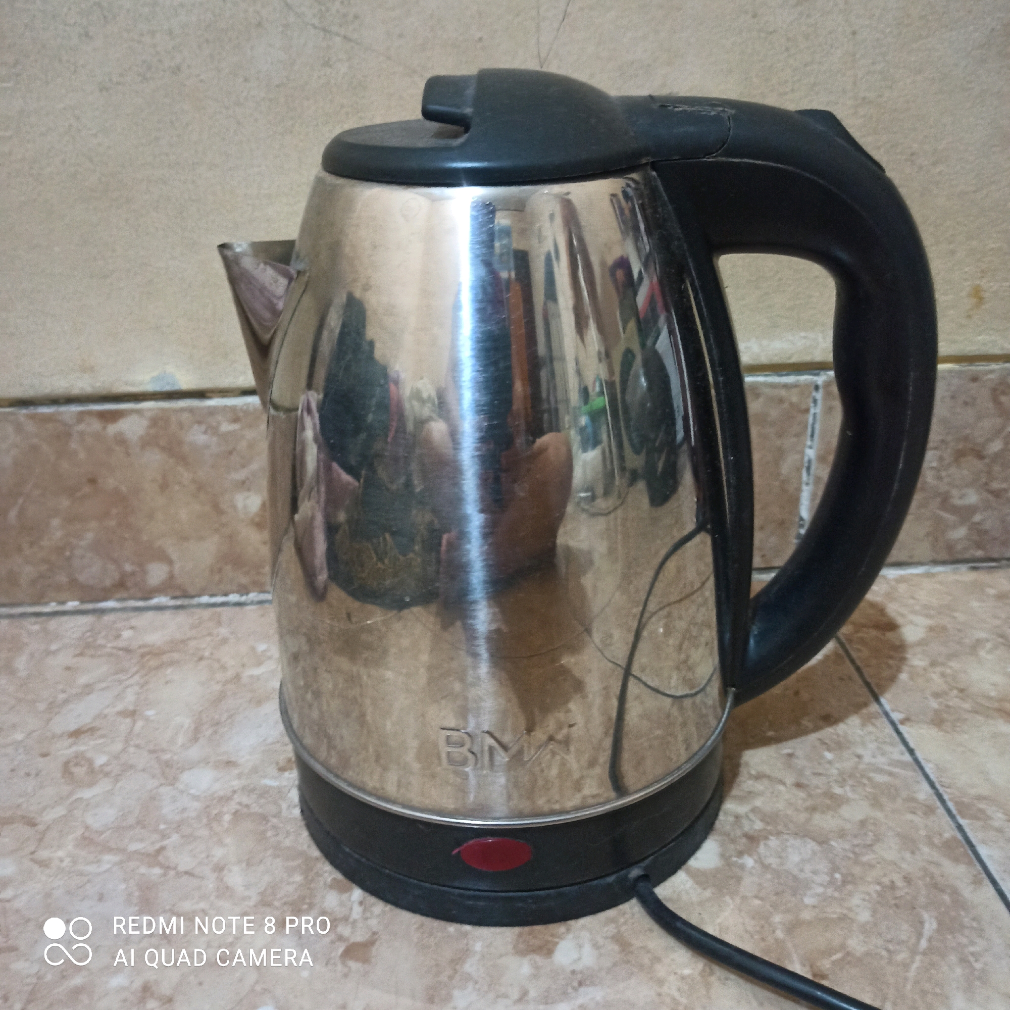 A stainless steel electric kettle with a black plastic handle and lid is positioned on a tiled surface. The kettle has a shiny, reflective exterior that shows distorted reflections of its surroundings, including what appears to be furniture and possibly people. A red indicator light is visible near the base of the kettle. The background consists of a textured wall. The image appears to be taken indoors, in what could be a residential kitchen or dining area, possibly in East Jakarta, Indonesia, given the presence of text overlay indicating "REDMI NOTE 8 PRO AI QUAD CAMERA". There are no people actively interacting with the kettle. The overall scene is static and domestic.