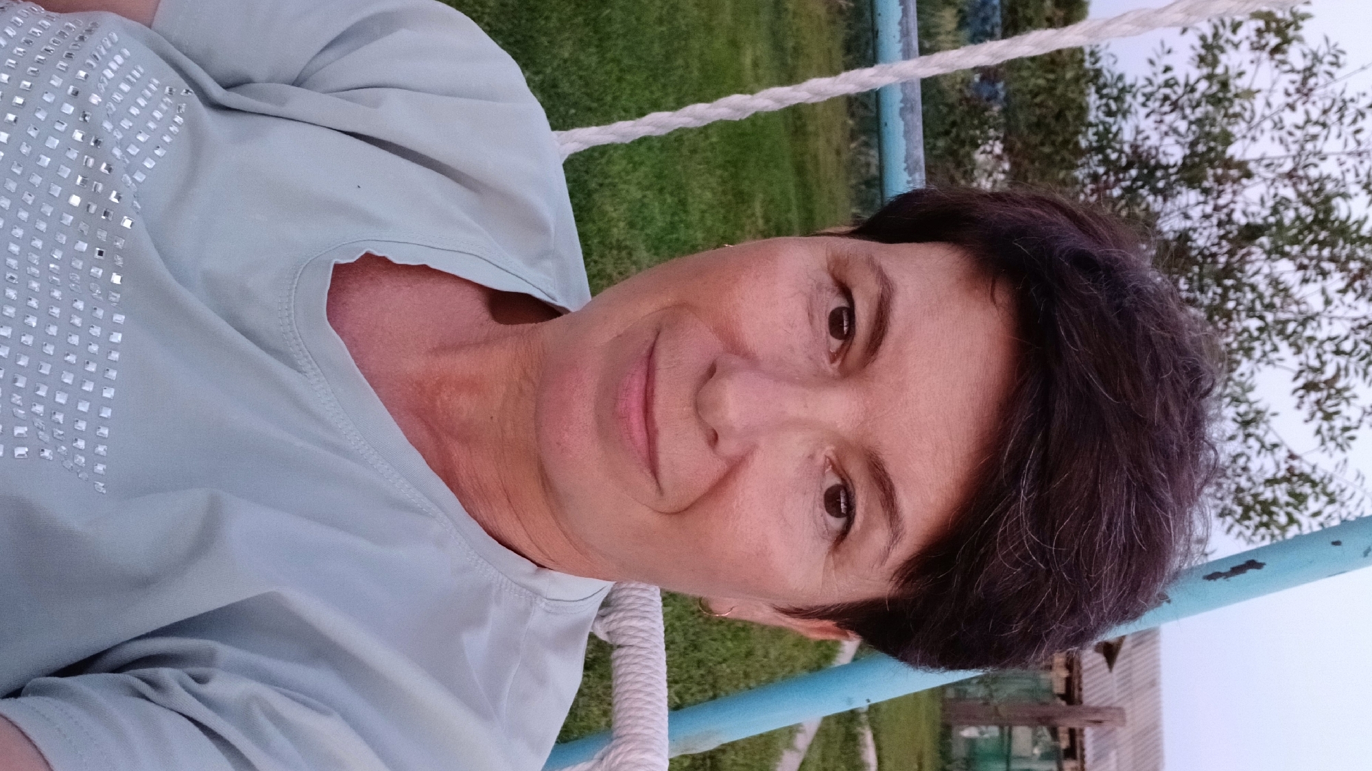 A woman with short, dark hair and fair skin is seated on a swing in an outdoor setting. She is wearing a light blue t-shirt adorned with silver rhinestones. The background features a green lawn, trees, and a rustic wooden structure. The lighting suggests it is either early morning or late evening. The woman is looking directly at the camera with a slight smile.