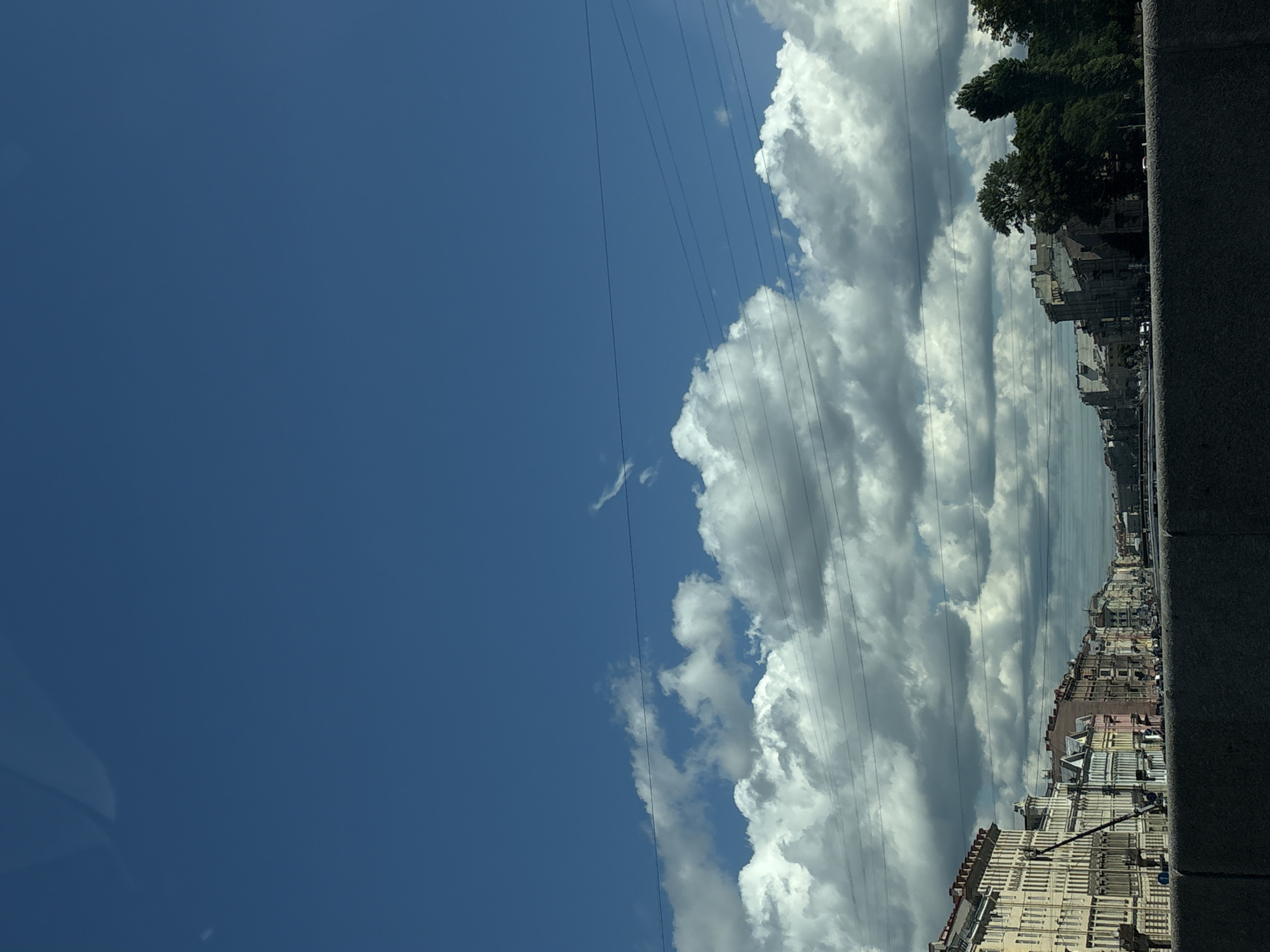 The image depicts a view of a canal in Saint Petersburg, Russia, with buildings lining the waterway. The sky is a bright blue with scattered white clouds, and several utility lines are visible against the sky. The buildings are multi-story, with varied architectural styles and colors. Some trees are present on the right side of the canal. The foreground shows a stone embankment. There are no people or discernible actions visible in the image.