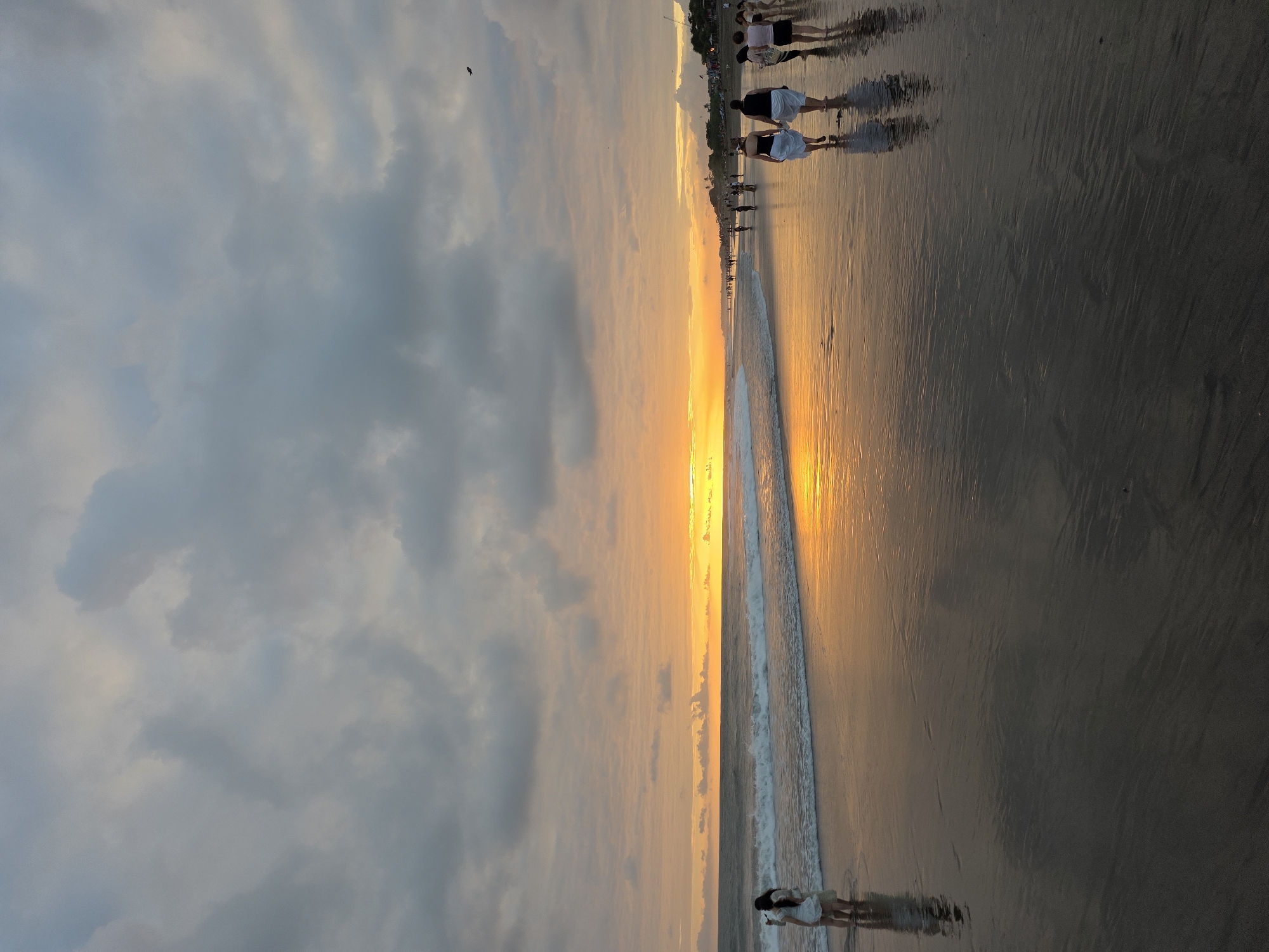 This is a vertical, eye-level, full shot of a beach at sunset. The sky is filled with dramatic, dark gray clouds with patches of bright orange and yellow light peeking through from the setting sun on the horizon. The ocean waves are gently rolling in, creating white foam on the wet sand. The wet sand reflects the colors of the sky and the sunset, creating a mirrored effect.
There are several people walking along the beach. On the left side of the frame, two women are standing in the shallow water, facing away from the camera. They are both wearing white dresses. Further down the beach, a group of people are walking in the same direction as the waves. They are silhouetted against the bright sunset. One person in the distance appears to be flying a kite. The location is a beach in the city of North Kuta, Indonesia.