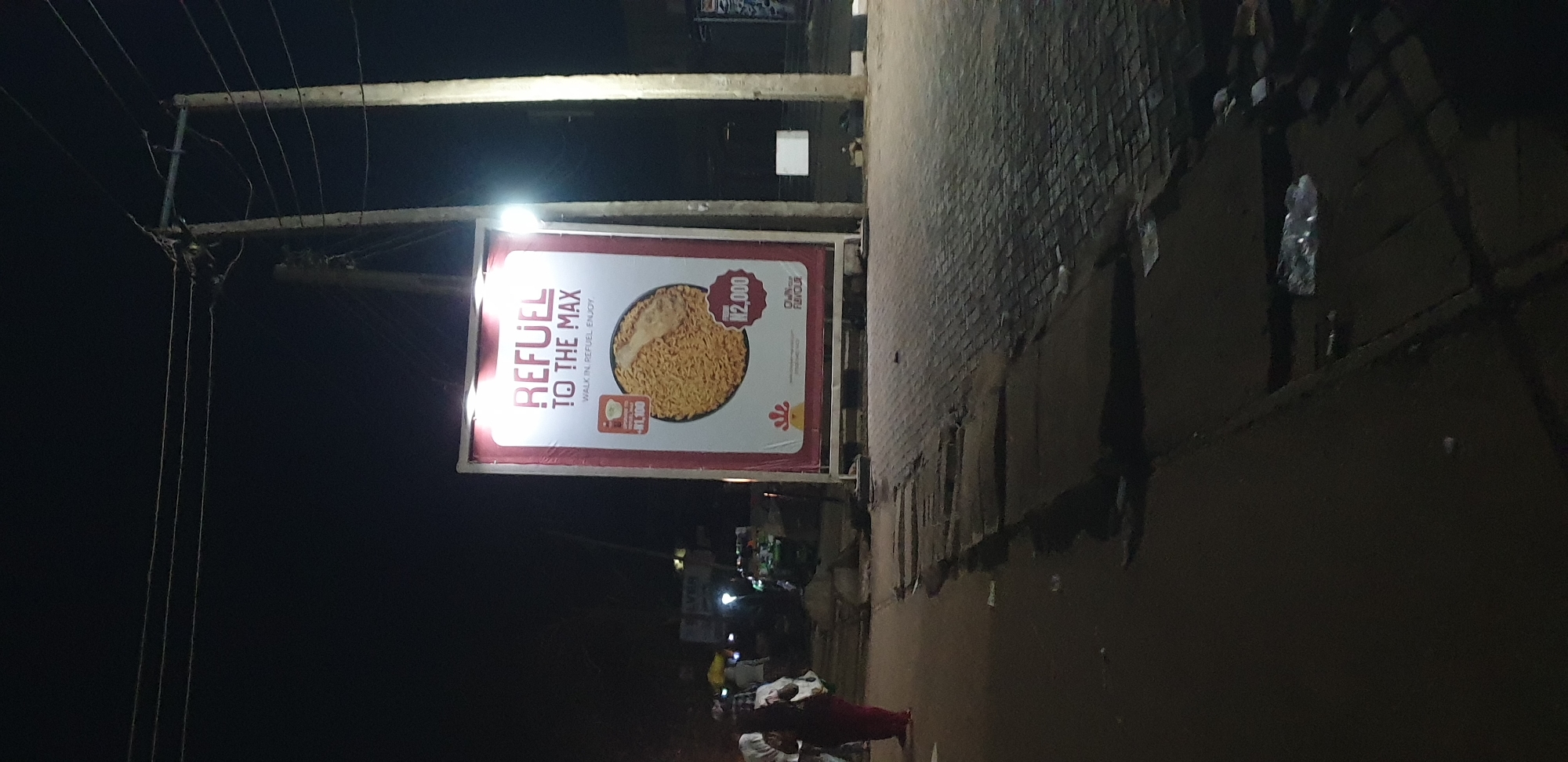 This is a nighttime photograph depicting a street scene in Sabon Tasha, Nigeria. The primary subject is a brightly illuminated outdoor billboard positioned centrally in the frame. The billboard has a red border and a white background, displaying the bold text "REFUEL TO THE MAX" and "WALK IN. REFUEL. ENJOY." It features a large circular image of a noodle dish, with an adjacent price tag indicating "₦2,000." A smaller image, possibly a cup of noodles, is shown with a price of "FROM ₦1,100." A red chicken-head logo and the text "OWN OUR FLAVOUR" are visible at the bottom of the billboard, along with a partial web address "chicken-republic.com." Two overhead lights illuminate the billboard from above.
To the immediate right of the billboard stands a tall concrete utility pole with an illuminated street light attached. Another, slightly shorter utility pole, possibly wooden or concrete, is visible further back and to the left of the billboard, supporting multiple overhead power lines.
In the foreground, the ground is dark and appears to be an uneven asphalt or packed earth surface. A visible drainage ditch or gully runs parallel to the billboard's base. To the right of the ditch and the billboard, the ground is paved with interlocking bricks or pavers. A clear plastic bottle is lying on the dark ground in the lower right section of the image.
On the left side of the frame, a person wearing a dark top and red or maroon trousers/skirt walks away from the camera, carrying a white bag over their left shoulder. In the blurred background to the far left, several indistinct figures are visible, suggesting other pedestrians or activity. Distant, faint lights from buildings or street vendors can be observed in the dark background beyond the people. The overall scene is dimly lit by artificial sources, primarily the billboard and the street light.