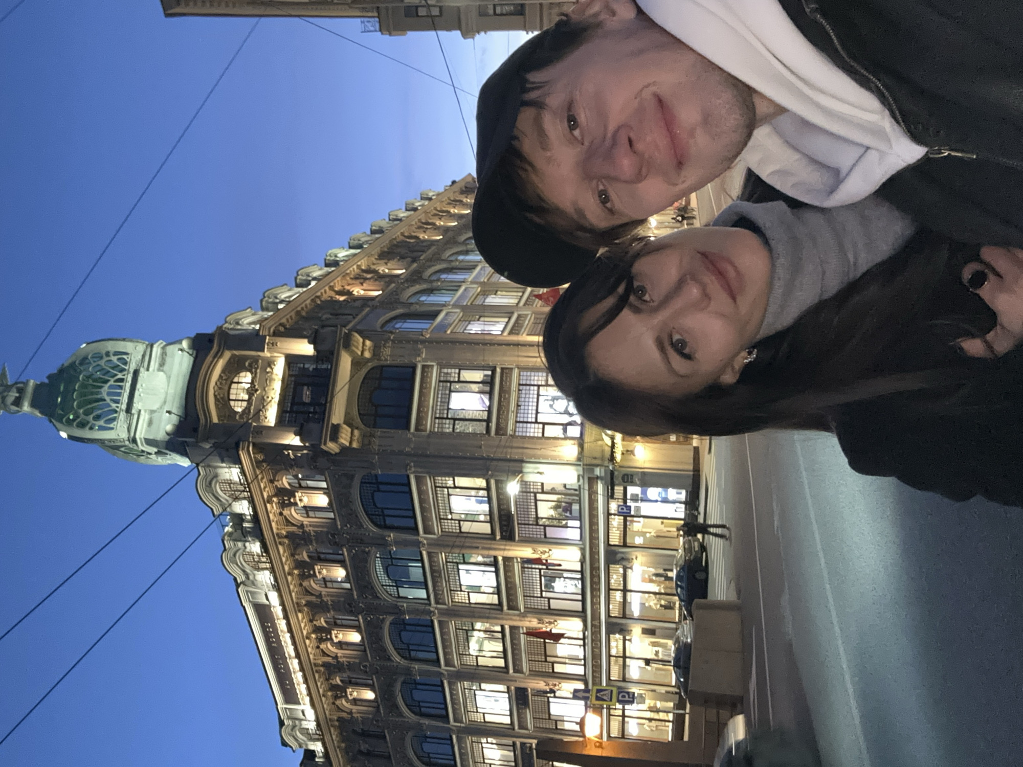 Two individuals are posing for a selfie in front of the historic Singer House building in Saint Petersburg, Russia. The building, notable for its Art Nouveau architectural style and domed tower, is illuminated with warm lighting against a deep blue evening sky. The selfie captures a portion of the street, cars, and pedestrian traffic, as well as utility poles and power lines. The building's facade features numerous windows and intricate architectural details.