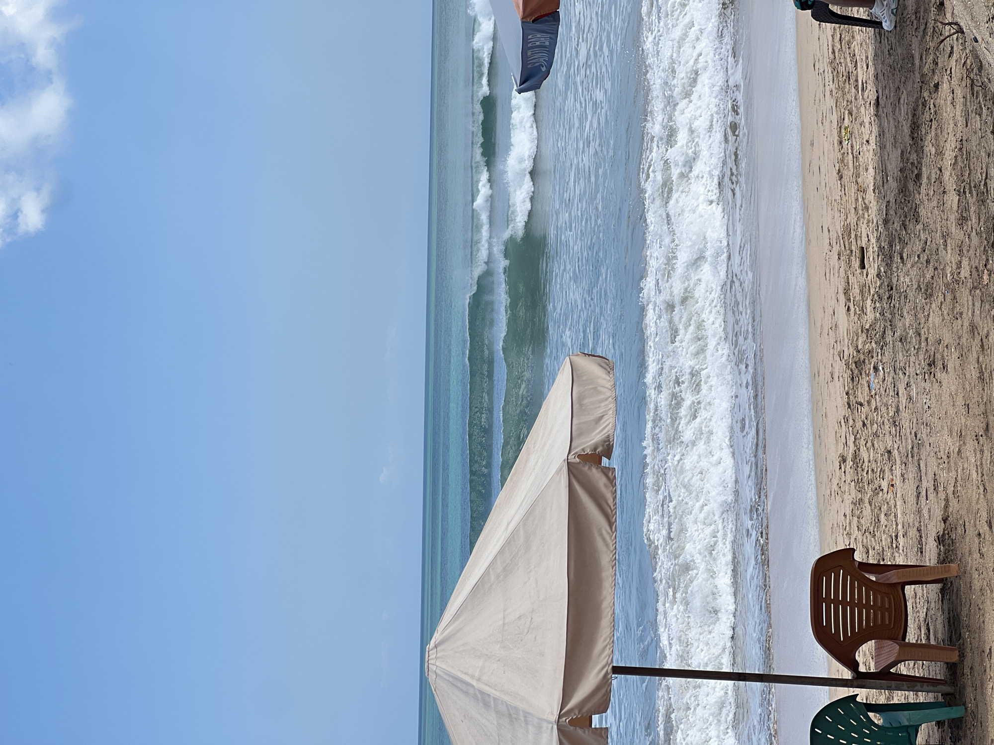 An outdoor, eye-level, full shot captures a beach scene in Kuta, Indonesia, with the ocean and sky dominating the frame. The sky is a bright, clear blue with a few wispy white clouds. In the foreground, the sandy beach is dotted with beach chairs. A large, light-colored beach umbrella shades two chairs, one green and one brown. To the right, another umbrella with an orange and blue canopy is visible. The ocean is a vibrant turquoise, with several sets of waves rolling towards the shore, some breaking into white foam. The water appears calm with visible currents and ripples. The sand is light brown, with a few pieces of debris scattered on it. The image evokes a serene and peaceful atmosphere, with the sound of the waves and the warmth of the sun.