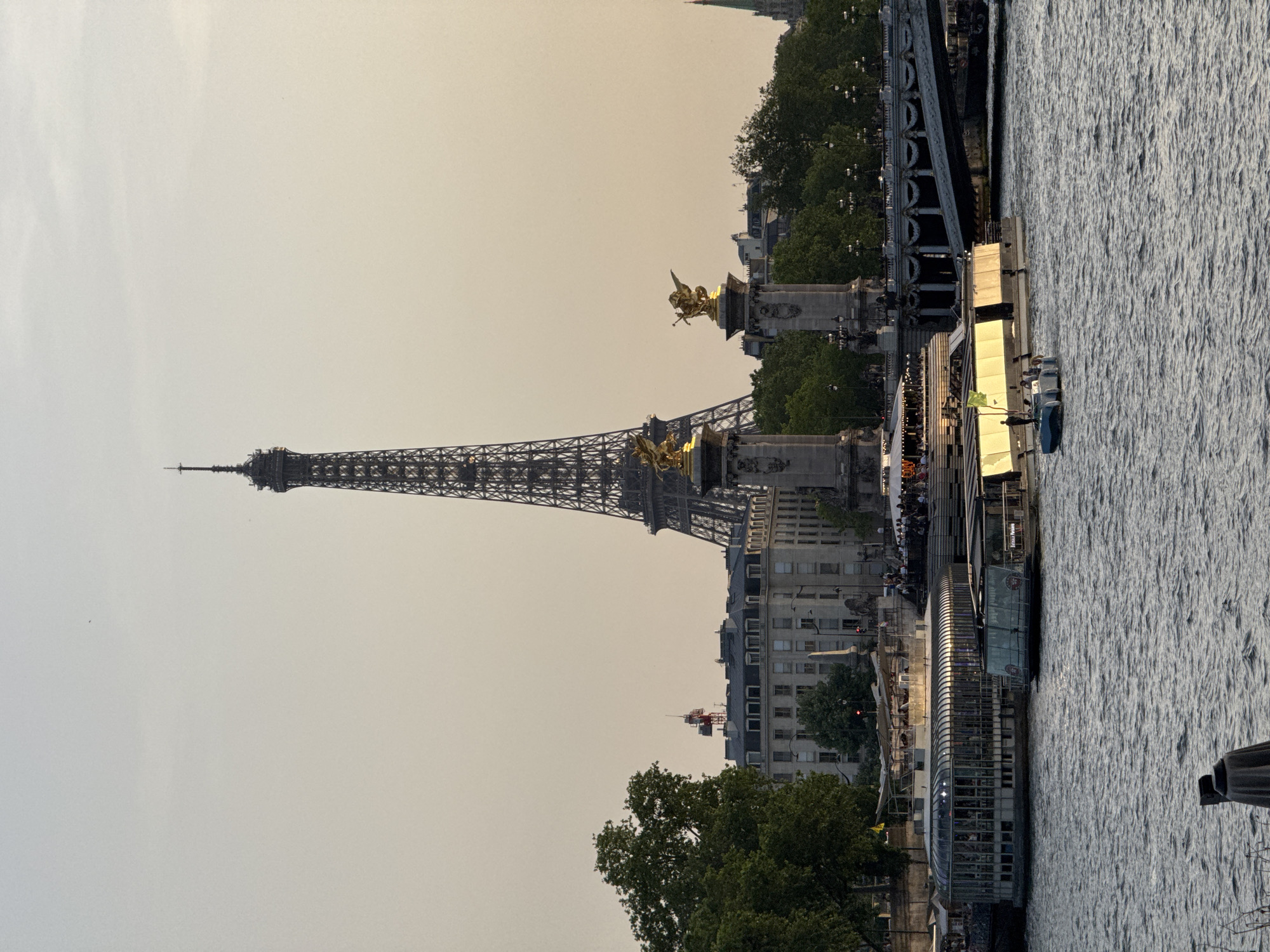 The image depicts a view in Paris, France. The Eiffel Tower is visible in the background. The Alexandre III Bridge is also present, with its golden statues and ornate design. The Seine River flows in the foreground. A riverboat or barge is docked along the riverbank, with people visible on board. Trees and buildings line the riverbank. The sky is overcast. A construction crane is visible behind the buildings.