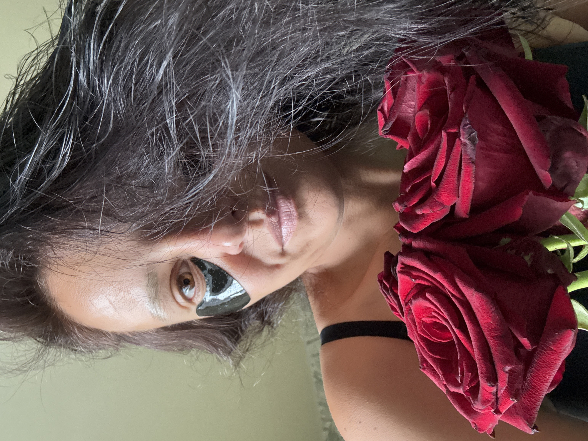 An angled, close-up portrait features an individual, likely female, with dark, textured hair showing prominent gray strands, suggesting natural aging or a styled look. A black, tear-shaped under-eye patch is applied below the visible brown eye, which gazes directly at the viewer. The person's lips are slightly parted, adorned with a light gloss. A thin black choker encircles the neck, with a matching black strap from clothing visible on the shoulder.
Two dark red roses with velvety petals are prominently placed next to the person's head, adding a touch of natural beauty and color to the composition. The background is a softly blurred, light-toned wall, indicating an indoor setting illuminated by what appears to be soft, natural daylight. The person's expression is neutral and direct. No text is visible. The image is associated with Chaikovsky, Russia.