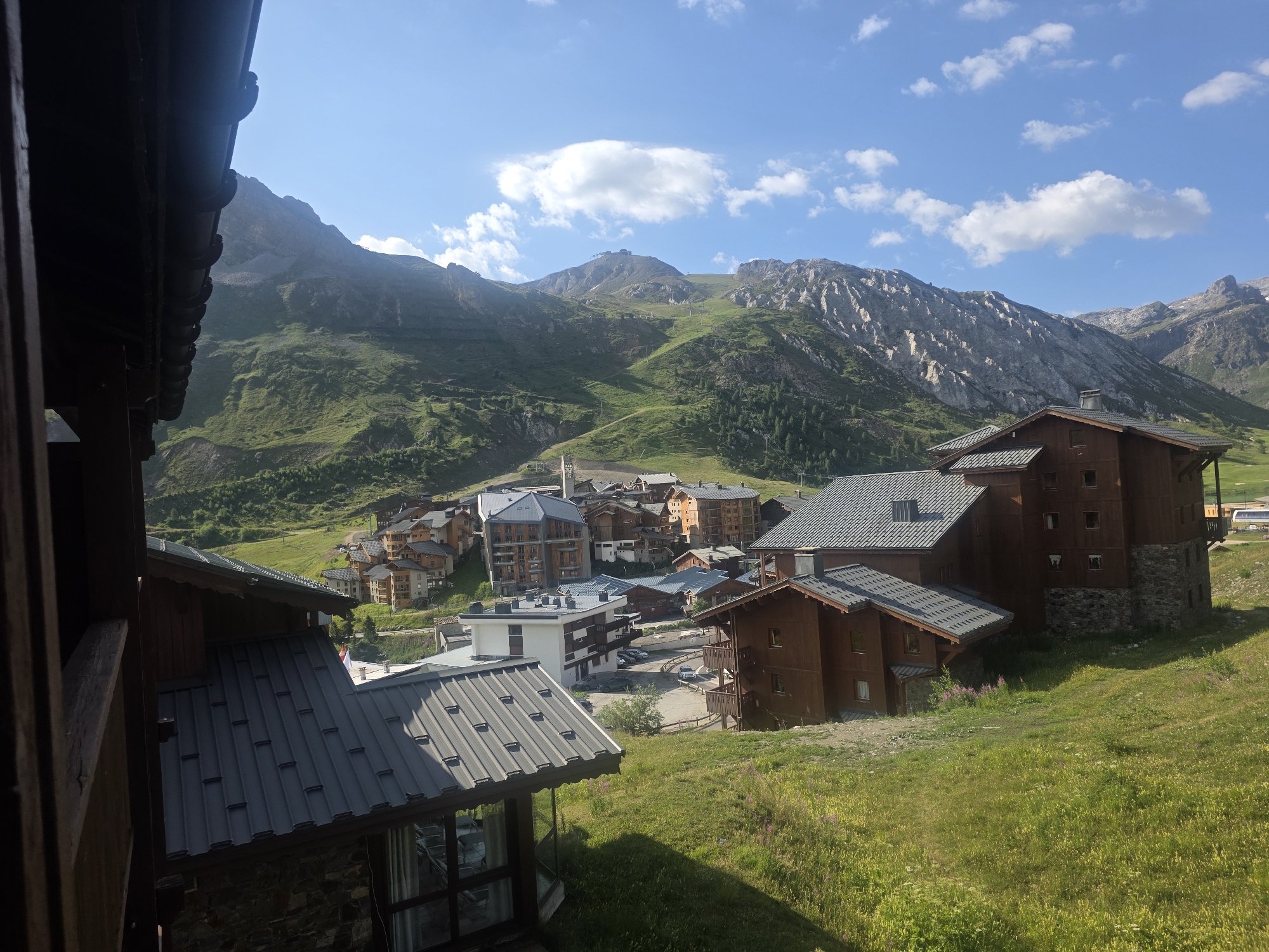 The image captures a scenic view of the Tignes Val Claret ski resort in Tignes, France, during daylight hours under a partly cloudy sky. The foreground features the dark wooden structure of a building with a dark metal roof, partially obscuring the view. In the mid-ground, several multi-story buildings with wooden facades and dark roofs are clustered on a grassy hillside, indicative of alpine architecture. Some of these buildings appear to be hotels or chalets, a common sight in ski resorts. A road with a few parked cars is visible between the buildings.
The background is dominated by the majestic Tignes mountains, with their steep, rocky slopes covered in patches of green grass and dotted with sparse trees. Ski lifts and tracks are discernible on the mountainside, suggesting the area is operational for winter sports. The overall impression is one of a tranquil mountain village set against a dramatic natural landscape. No people are visible in the frame.