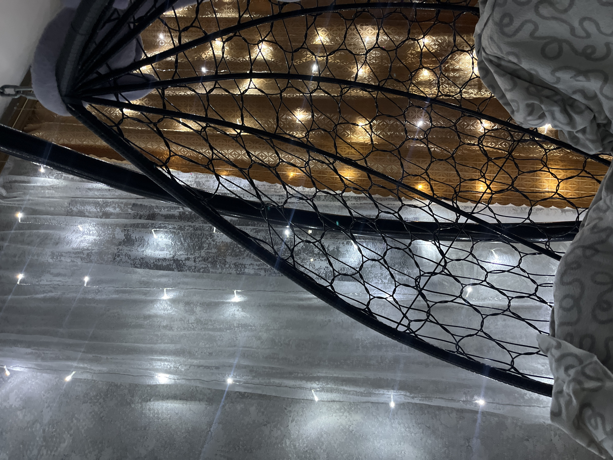 The image depicts a close-up view of a black metal frame with a net pattern, possibly part of a hanging chair or similar structure. The background consists of two distinct areas: the upper portion is filled with a soft, warm, golden glow from strings of lights against a textured surface, while the lower portion shows a series of gray steps with faint white lights draped along them. A portion of a patterned gray-and-white fabric is visible on the right side of the frame. The image appears to be taken indoors. The location of Novopokrovskaya, Russia, is not definitively indicated in the visual details of the image.