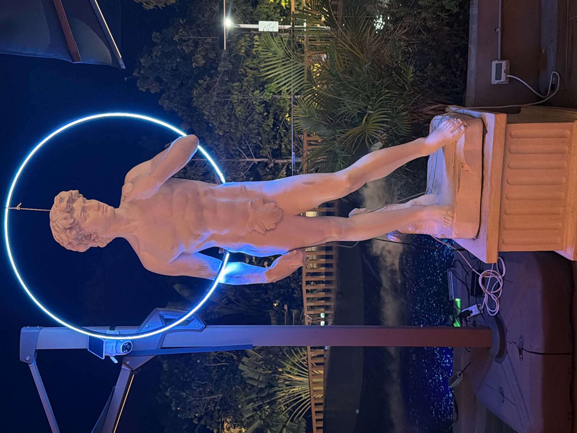 This media file features a statue of a male figure, resembling classical sculpture, in a dynamic pose. The statue is illuminated by a bright blue neon circle that encircles its torso and extends to its arms, creating a dramatic visual effect. The statue is positioned on a pedestal, which is part of a larger, multi-layered structure.
The scene is set outdoors at night, with the background revealing lush tropical foliage, including palm fronds, and a dark sky. Wooden lattice structures and lights can be seen in the distance, suggesting an outdoor venue or garden. The overall lighting is low, with the neon blue light from the circle being the most prominent illumination source, casting a cool glow on the statue. Some wires and electrical components are visible near the base of the statue and pedestal, including a power outlet with a green light on.
The image captures a sense of artistic display or installation, possibly in a resort, public space, or gallery setting in Ko Samui District, Thailand. The combination of classical art and modern neon lighting creates a striking juxtaposition. No visible text is present in the image.