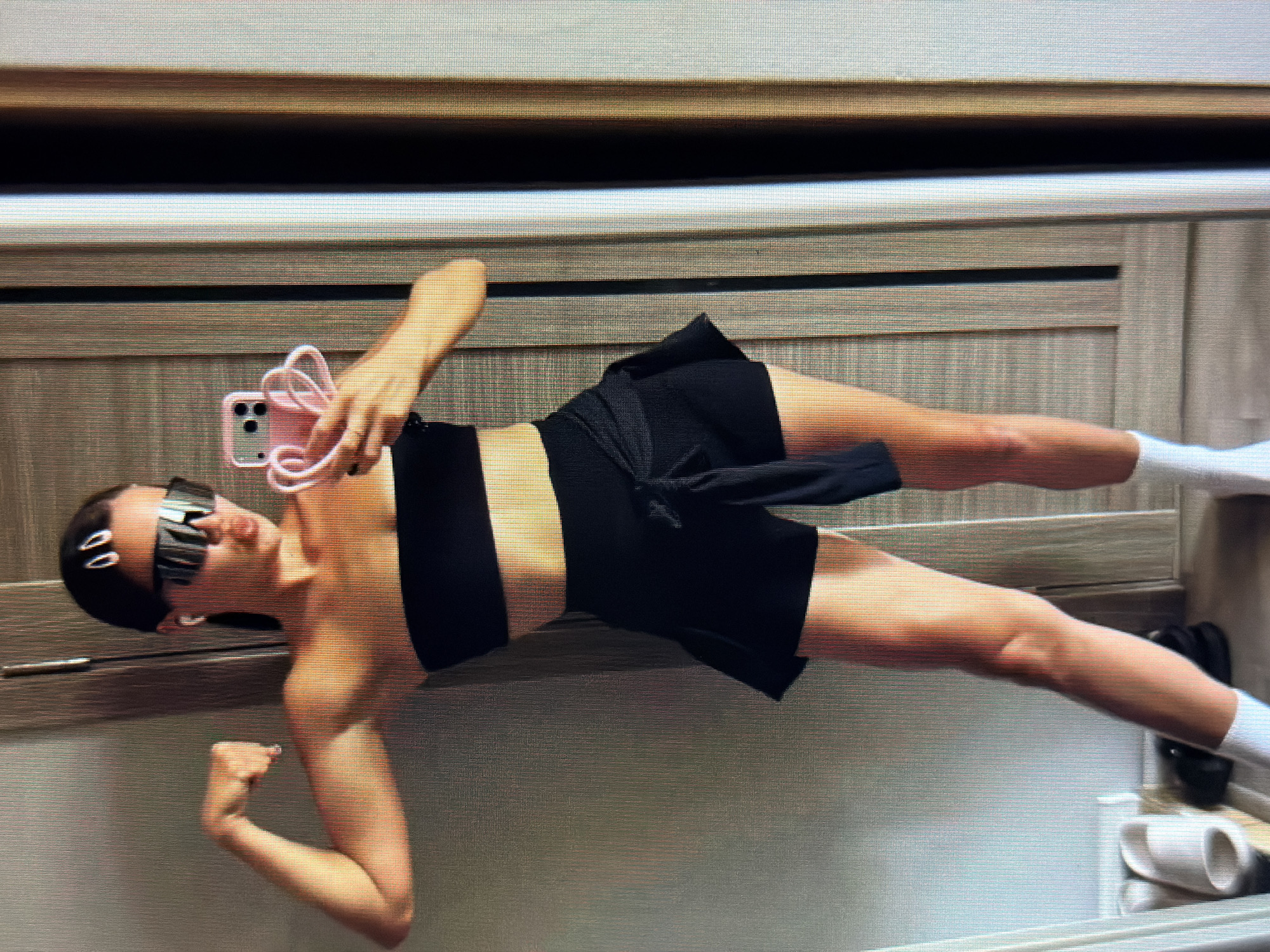 A woman is standing in what appears to be a room with wood-paneled walls, possibly a hallway or a dressing area. She is looking into a mirror and taking a selfie with her phone. She is wearing a black strapless top, black shorts with a tie waist, white socks, and white sneakers. She also has on large, futuristic-looking sunglasses and white hair clips. Her left arm is flexed, showing off her bicep. The background shows a door and some shelving with footwear. The lighting suggests it is daytime, and the overall setting is casual and personal. There is no indication in the image that this scene is located in the city of Sirius, Russia.