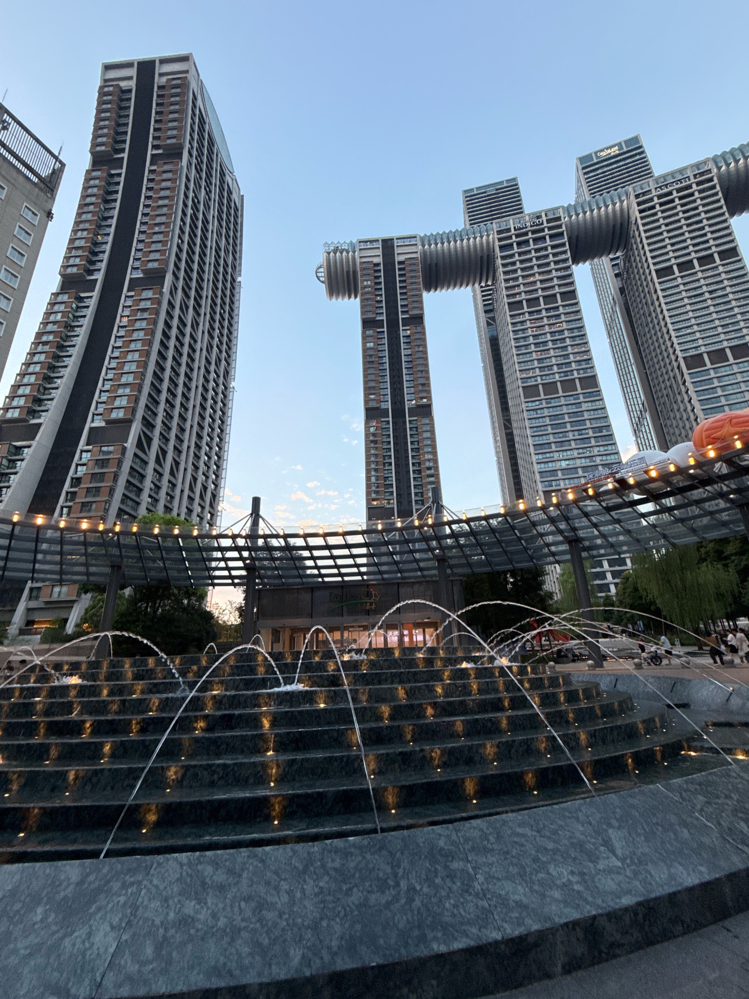 The image depicts a tiered fountain in the foreground, with water jets arcing over the dark, stone steps, each illuminated by a small, warm light. In the background, a row of modern skyscrapers dominates the cityscape. One particularly striking building features a unique design where two sections are connected by a horizontal bridge-like structure. The sky above is a clear, pale blue, suggesting it's either early morning or late afternoon. The scene appears to be an urban public space, possibly a plaza or park area adjacent to a commercial or residential complex. The location is specified as the Yuzhong District, China. There are several people visible in the background, some sitting, others standing, suggesting a public gathering place.
