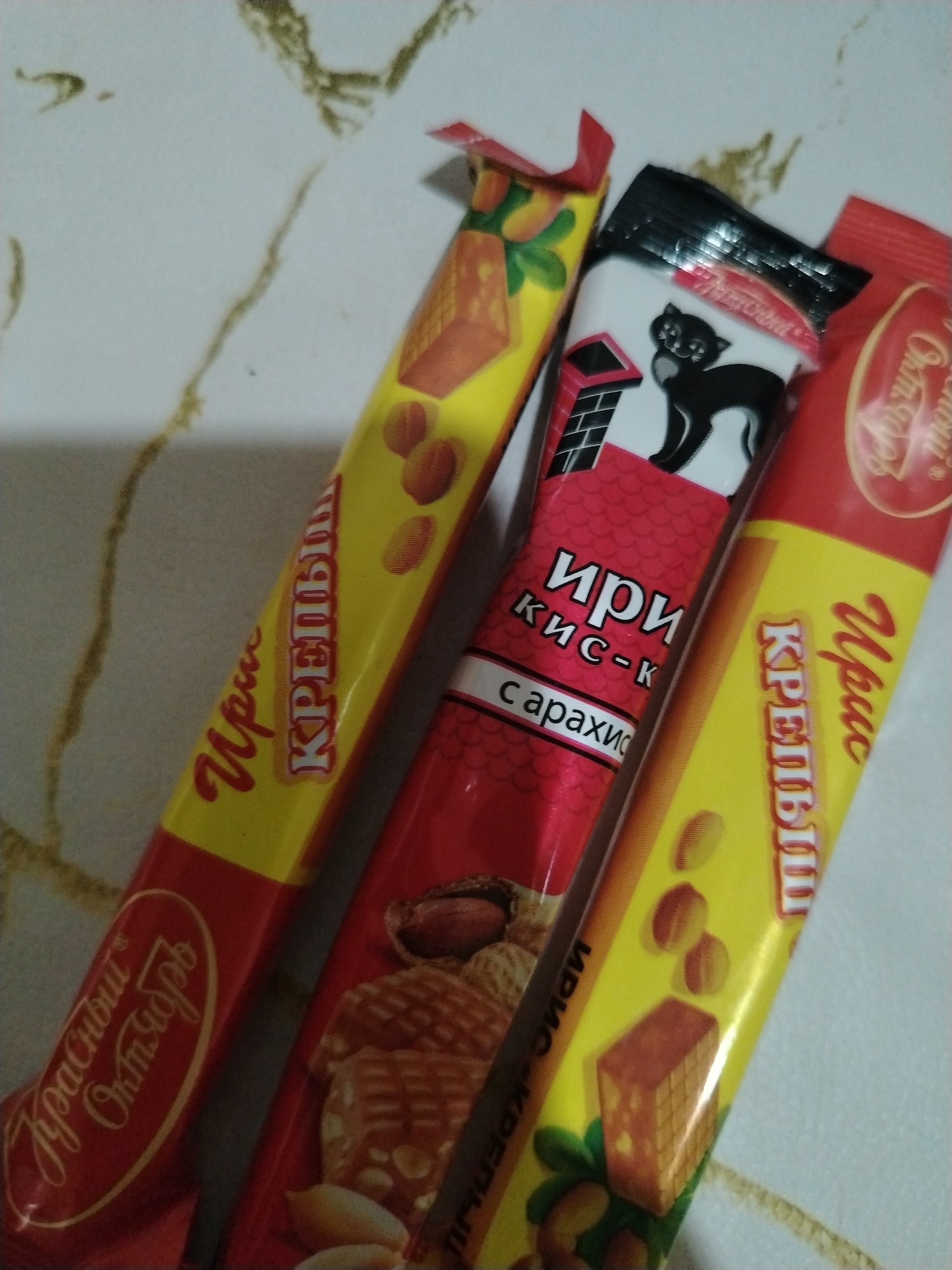 The image shows three candy bars lying on a light-colored surface with a marble-like pattern. The candy bars are predominantly red and yellow with text in Russian. One candy bar has a black cat on the wrapper. The candy bars appear to be varieties of "Iris," possibly with peanuts. The brand name "Krasny Oktyabr" is visible, suggesting the product is manufactured by the Krasny Oktyabr confectionery factory, located in Moscow, Russia. The image does not provide enough information to determine if the location is Rusanovo, Russia.