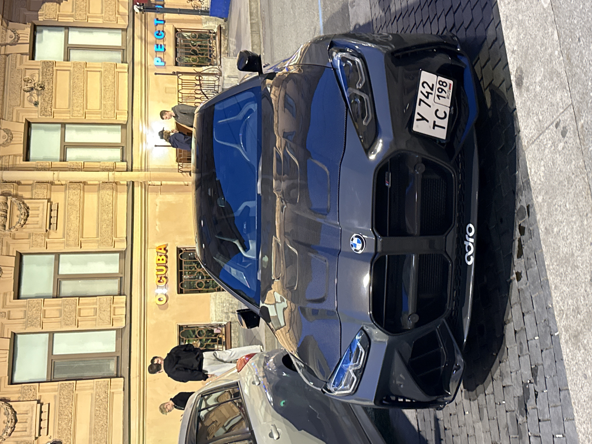 A dark grey BMW M4 coupe is parked on a cobblestone street in front of a building in Saint Petersburg, Russia. The car has a carbon fiber hood and a license plate that reads "Y 742 TC 198 RUS". The building behind the car is light yellow and has windows with decorative molding. There are two people standing on the sidewalk in front of the building, one wearing a black hoodie and grey pants, and another person in a black hoodie. There are also two people standing near the building entrance, one in a dark jacket and the other in a grey hoodie. One of the people is holding something to their mouth. A silver car is parked to the left of the BMW. Signs with Russian text are visible on the building facade.
