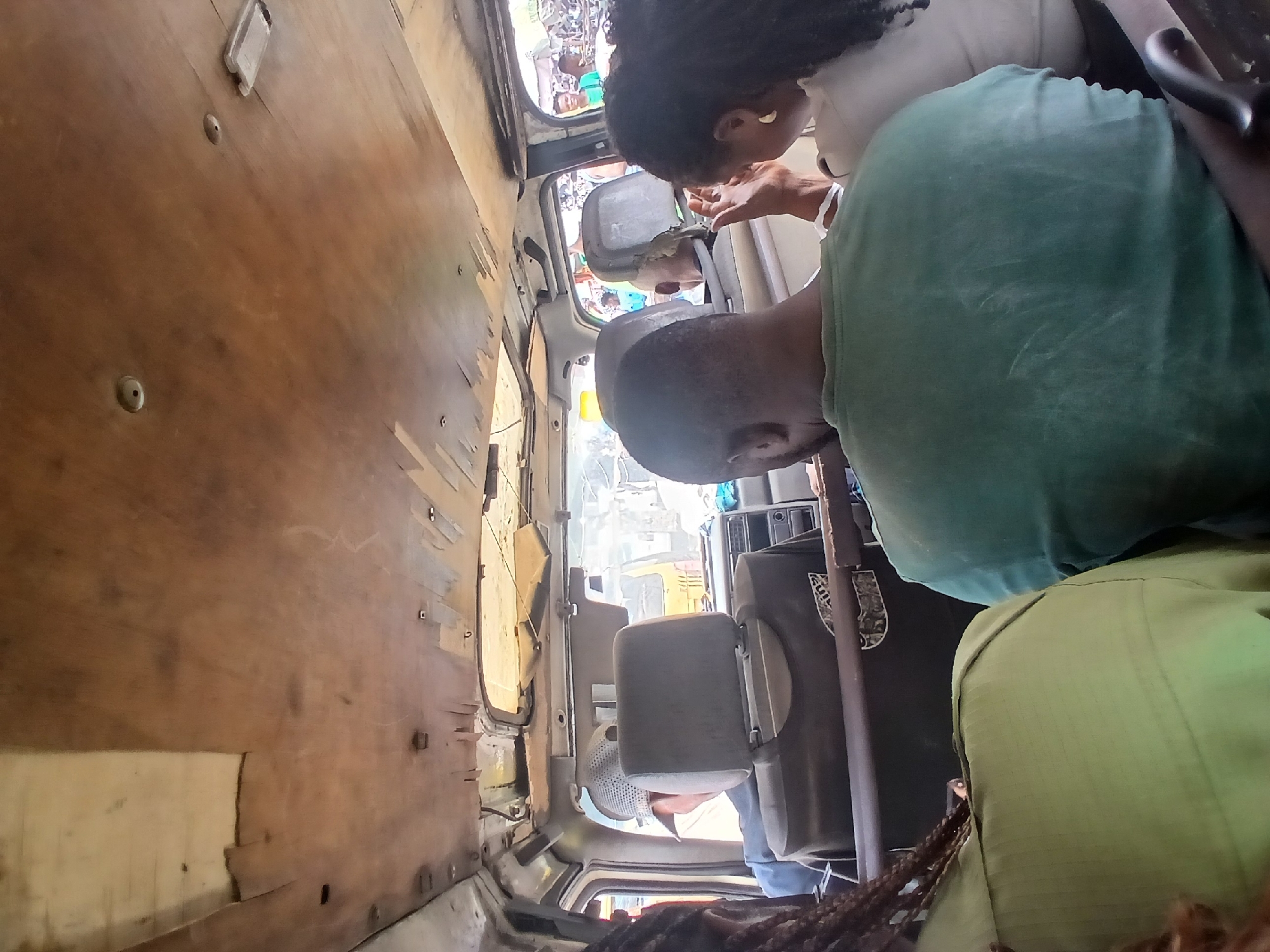 The interior of a crowded van is captured from a rear-facing perspective, looking towards the front and out the windows. The van's ceiling is made of worn, wooden panels, some of which are peeling and damaged. Multiple people are seated in the van, with their backs or sides to the camera. In the foreground, a person in a green shirt is seated to the left, and another person in a similar green garment is to the right, partially obscured. In the middle ground, a man in a green t-shirt is visible from the back, and next to him, a woman with dark curly hair and a light-colored top is seen from the side, with her right hand raised. Further forward, a man wearing a cap and a blue jacket is seated, facing forward. Through the front windshield and side windows, a bustling outdoor environment with other vehicles, including a yellow van, and people can be seen. The scene appears to be within a city, potentially in Aganiyin, Nigeria, given the context provided, suggesting a busy public transportation scenario.