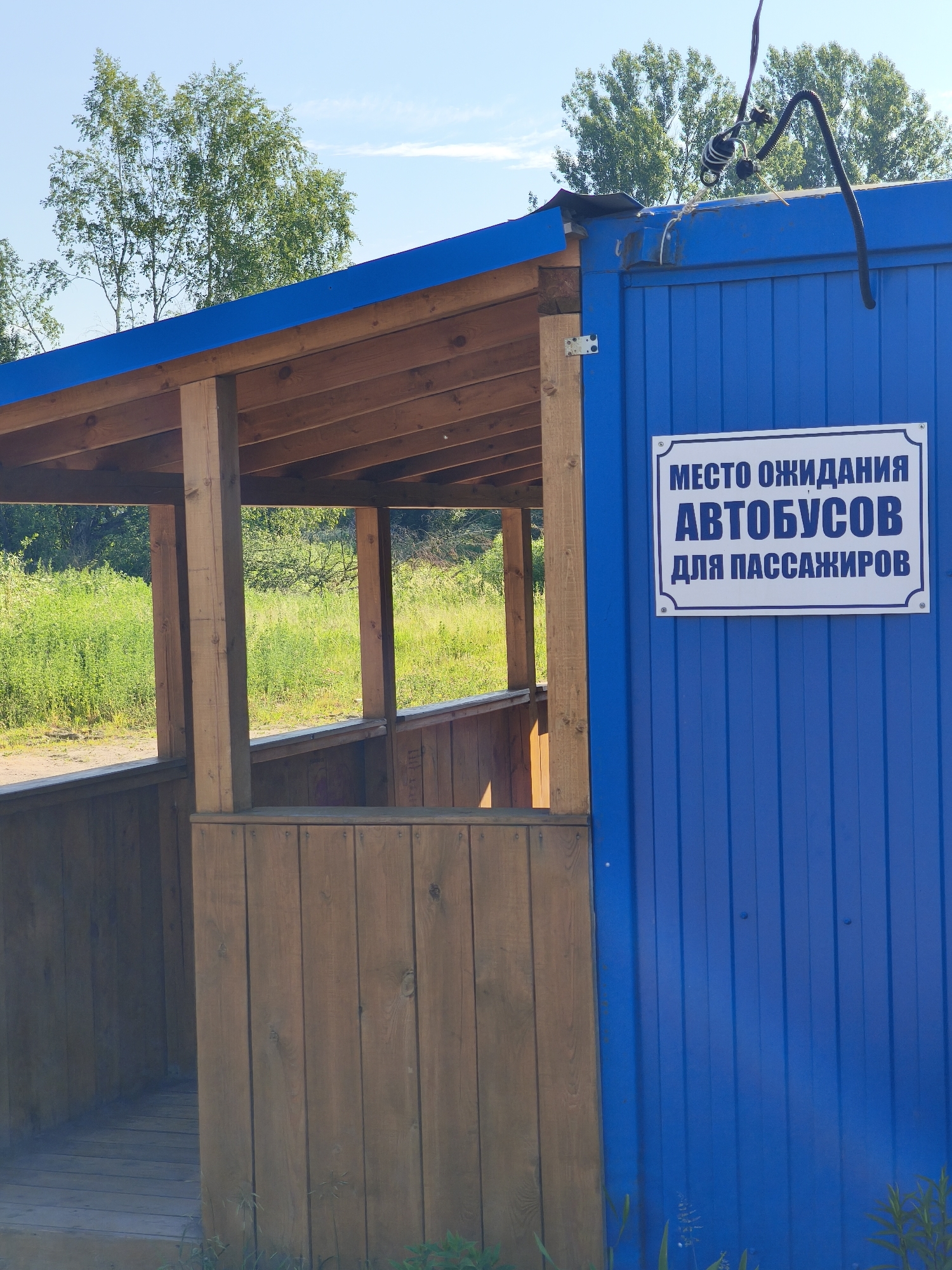 The image shows an outdoor bus stop shelter. The shelter has a wooden roof and posts, with a wooden floor and sides. A blue corrugated metal wall of a building forms the right side of the shelter. On this blue wall, there is a rectangular sign with a white background and blue text. The sign reads: "МЕСТО ОЖИДАНИЯ АВТОБУСОВ ДЛЯ ПАССАЖИРОВ", which translates to "BUS WAITING AREA FOR PASSENGERS". The bus stop appears to be located in a rural or semi-rural area, with green vegetation and trees visible in the background. The sky is clear and blue. The location is identified as Kady, Russia.